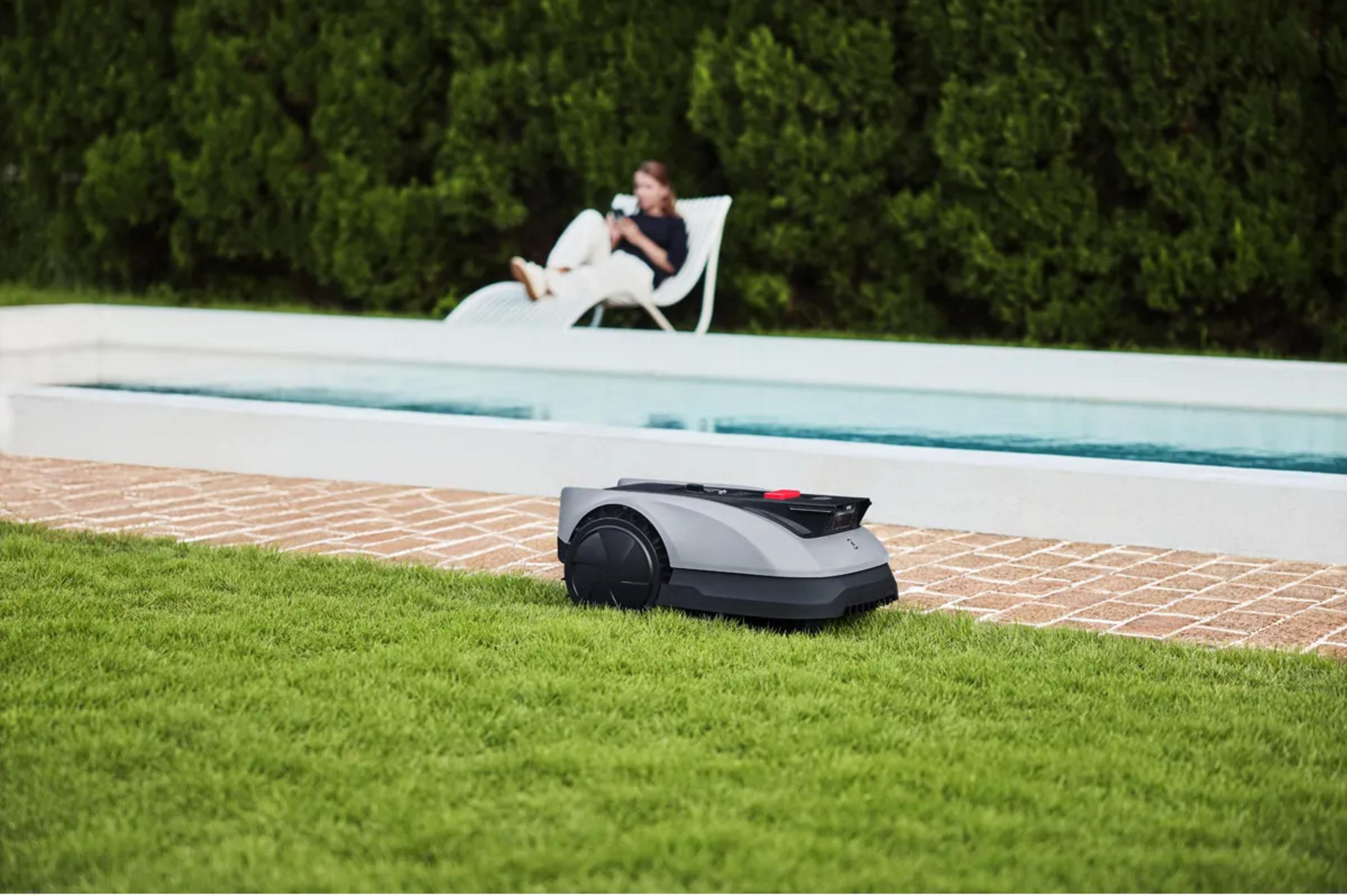 Ein Mähroboter steht auf einem Rasen neben einem Pool. Eine Frau entspannt sich im Hintergrund.