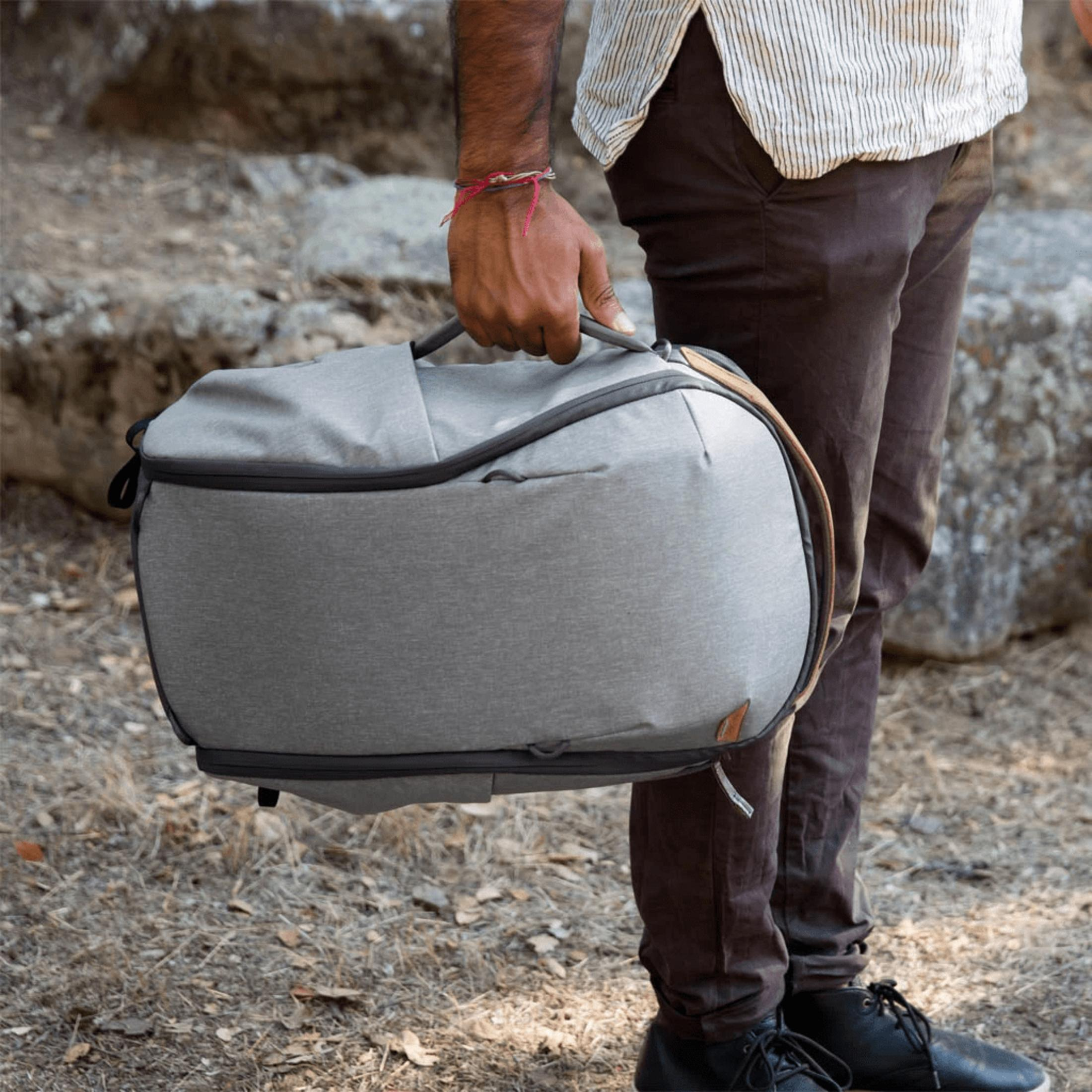 Un uomo tiene una borsa grigia, indossa pantaloni marroni e una camicia a righe.