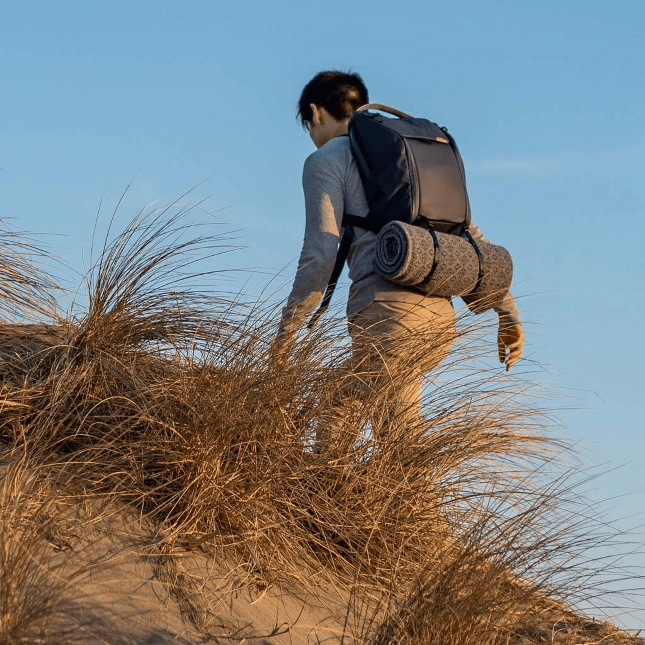 Ein Wanderer mit Rucksack klettert über eine sandige Düne mit trockenem Gras.