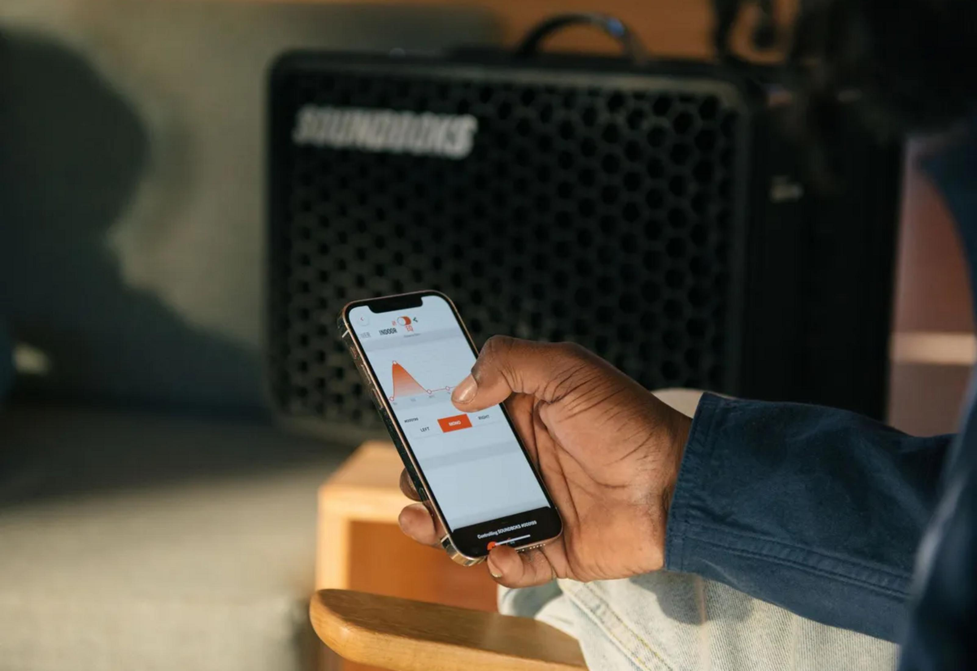 Eine Hand hält ein Smartphone mit einer EQ-App und steuert einen schwarzen Lautsprecher im Hintergrund.