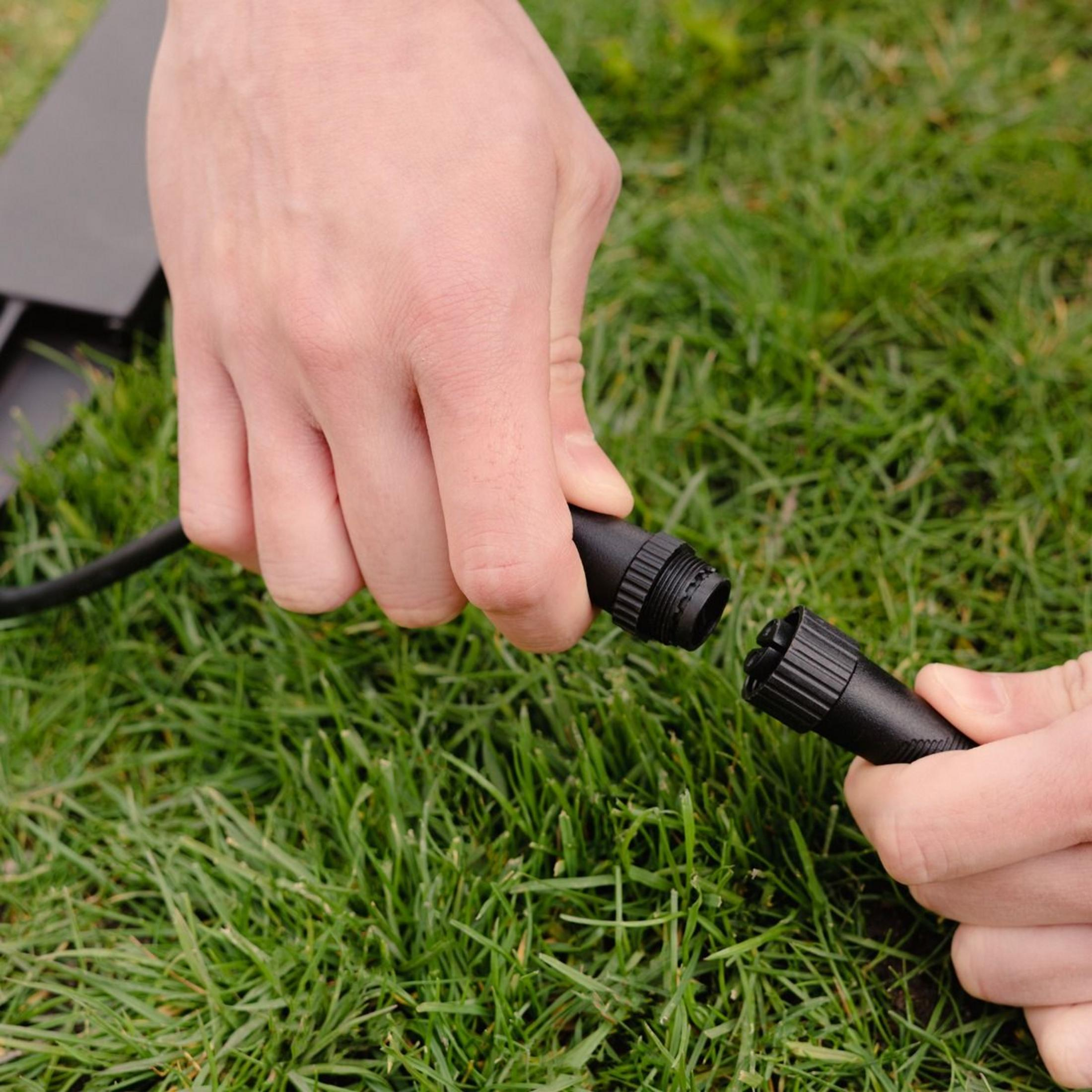 Hände verbinden ein schwarzes Kabel mit Steckern auf grünem Gras.