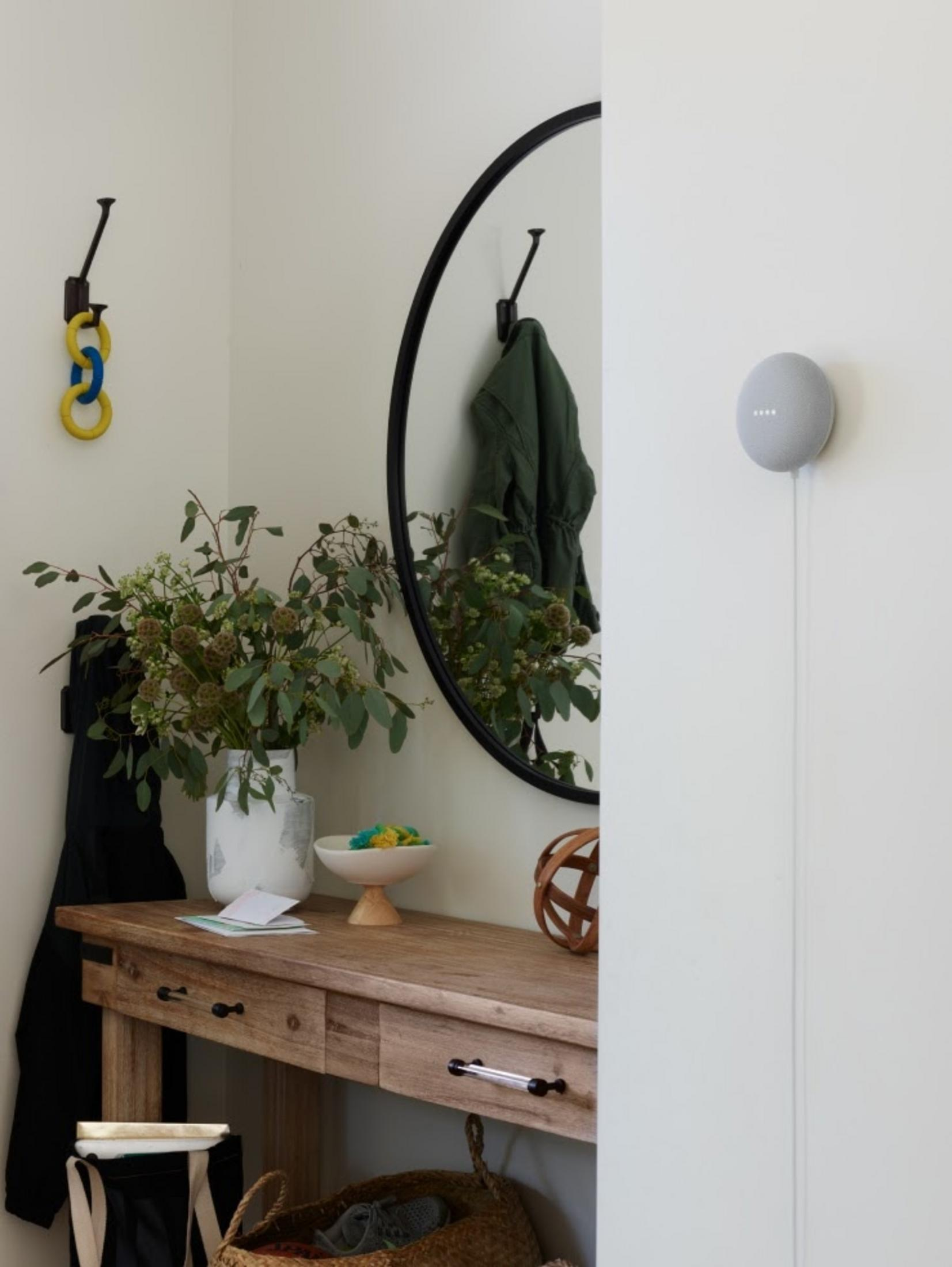 Un recibidor sencillo con mesa de madera, espejo, plantas y un Google Nest Mini gris.