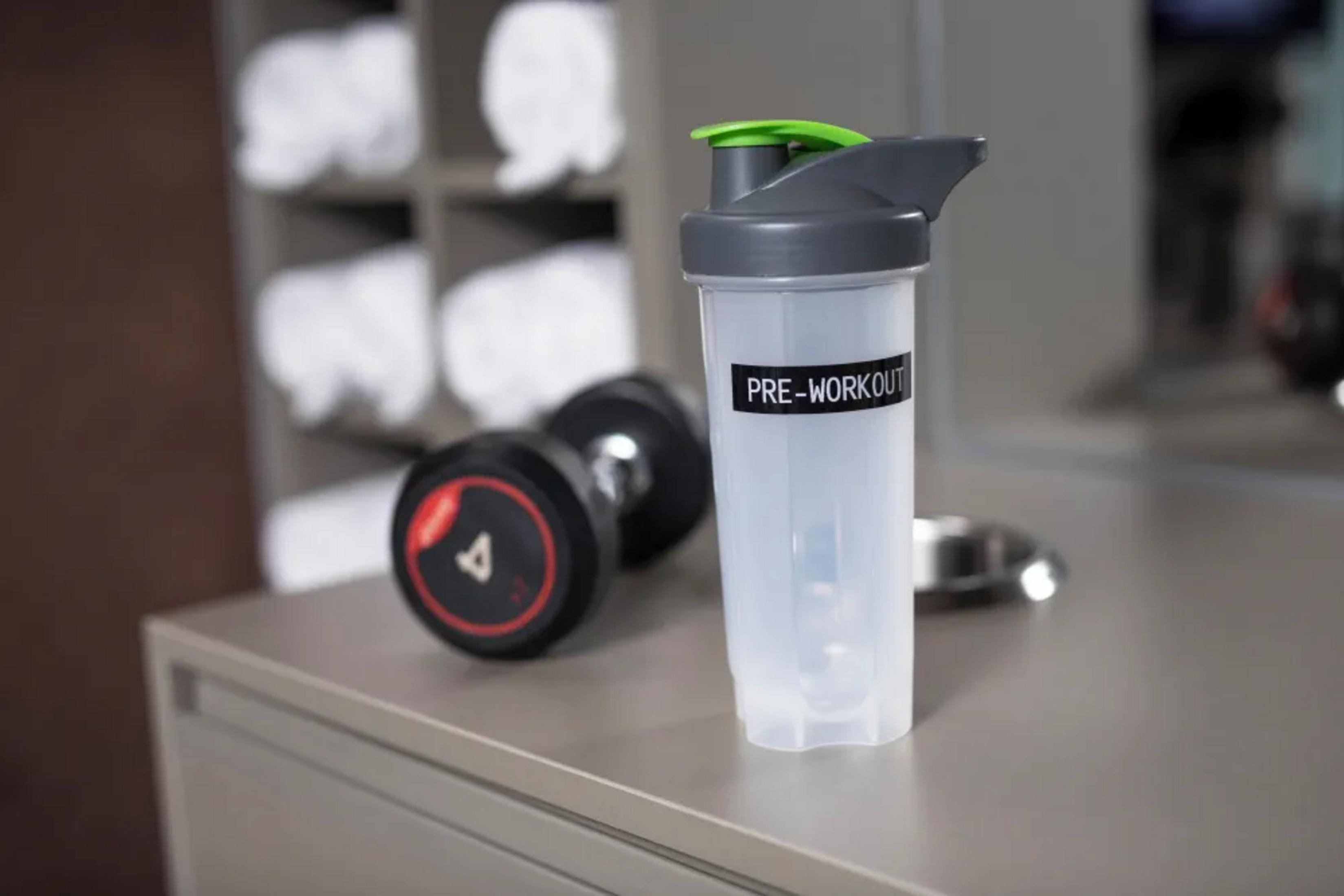 Ein Pre-Workout-Shaker mit Gewichten und Handtüchern im Hintergrund.