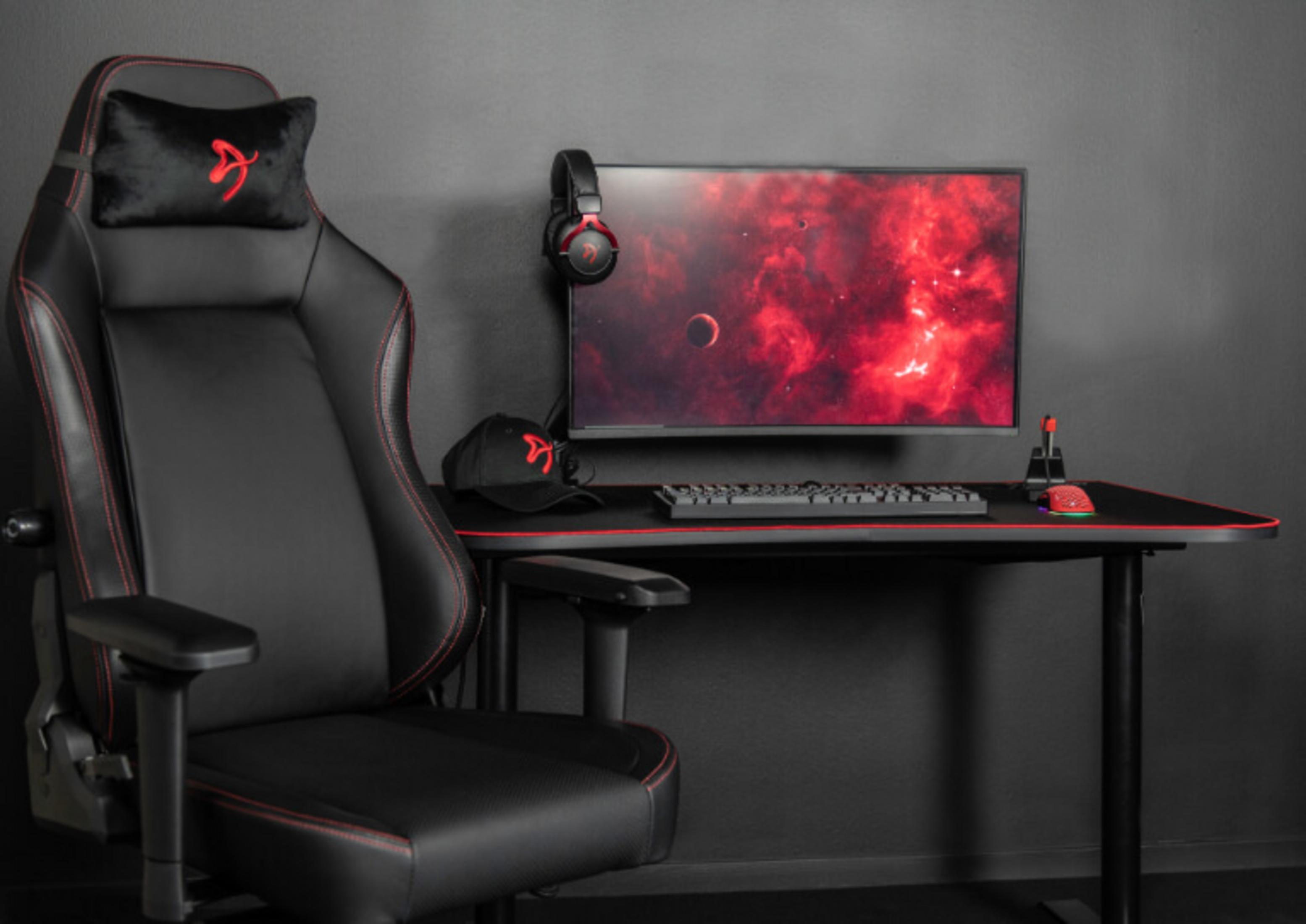 Una sedia da gaming nera con cuciture rosse, scrivania con monitor, tastiera, mouse e cuffie.