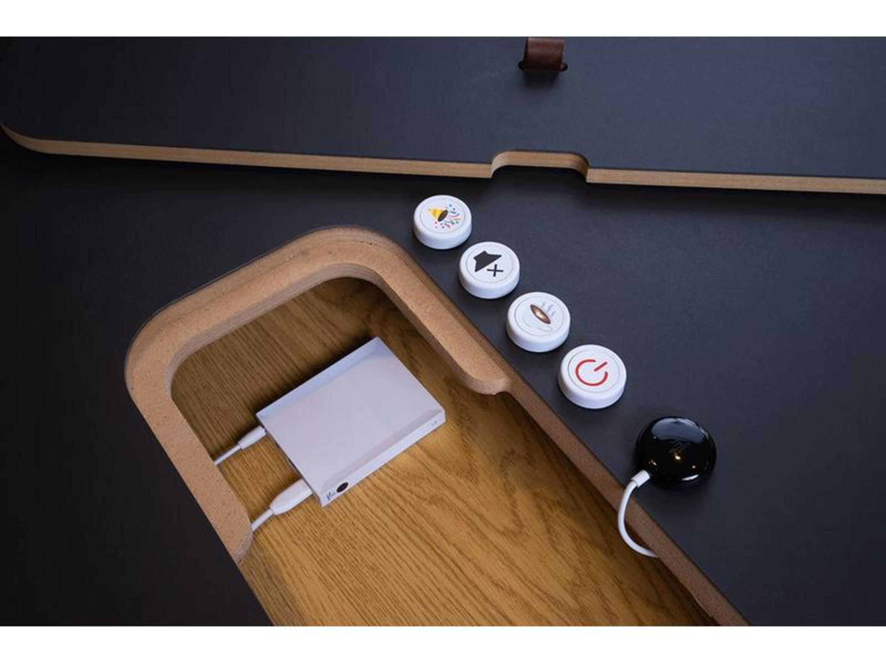 Weiße Powerbank mit Kabeln auf Holz, neben vier runden Knöpfen und einer schwarzen Kugel auf dunkler Oberfläche.