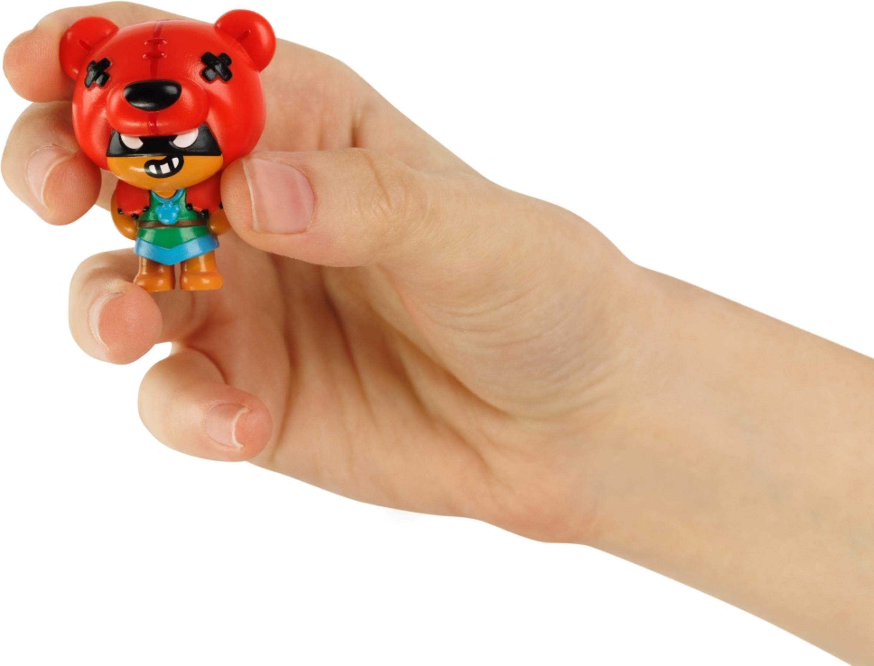 Une main tient une petite figurine d'ours rouge avec un masque noir et des vêtements verts.