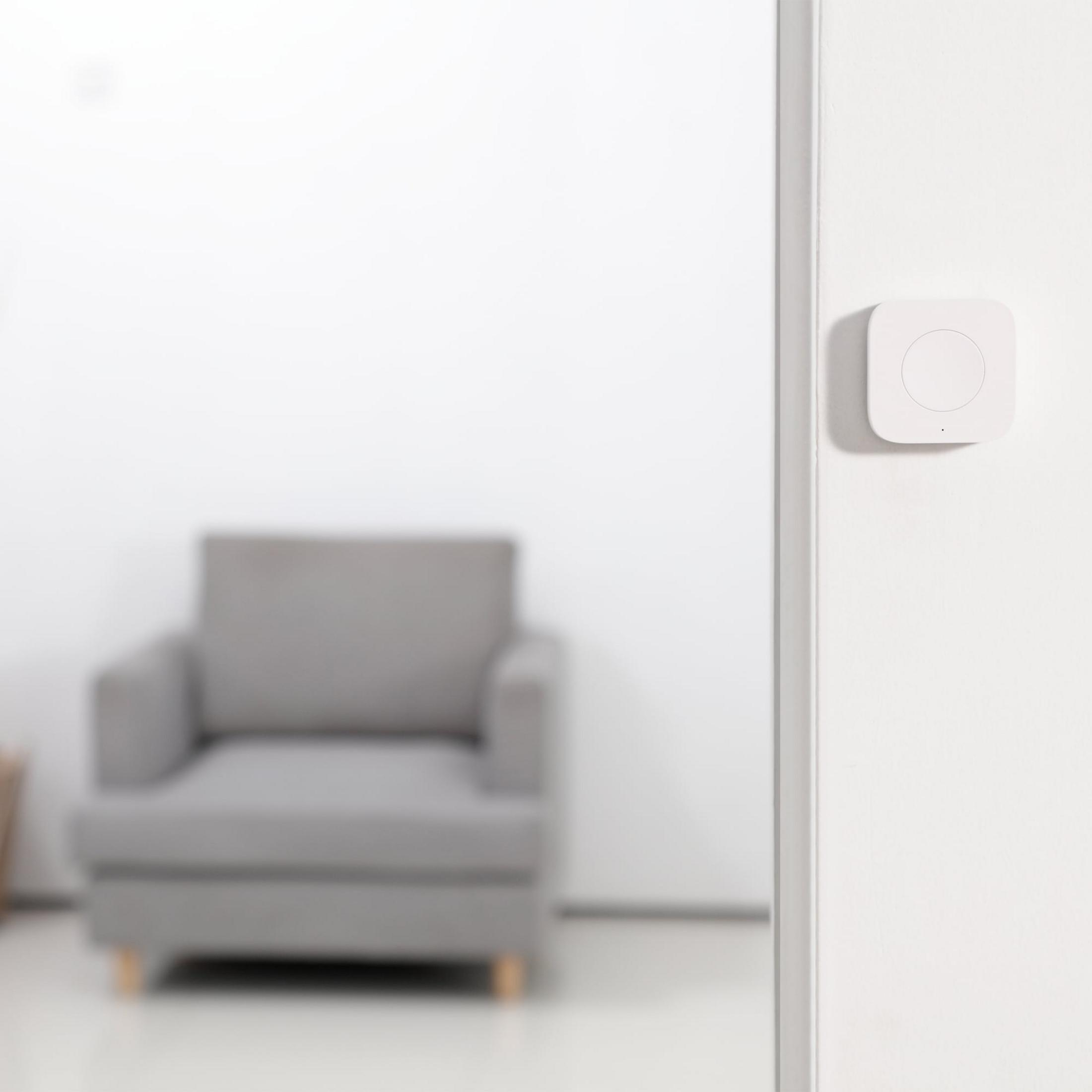 Un dispositivo quadrato bianco con un cerchio su un muro bianco, una poltrona grigia visibile dietro.
