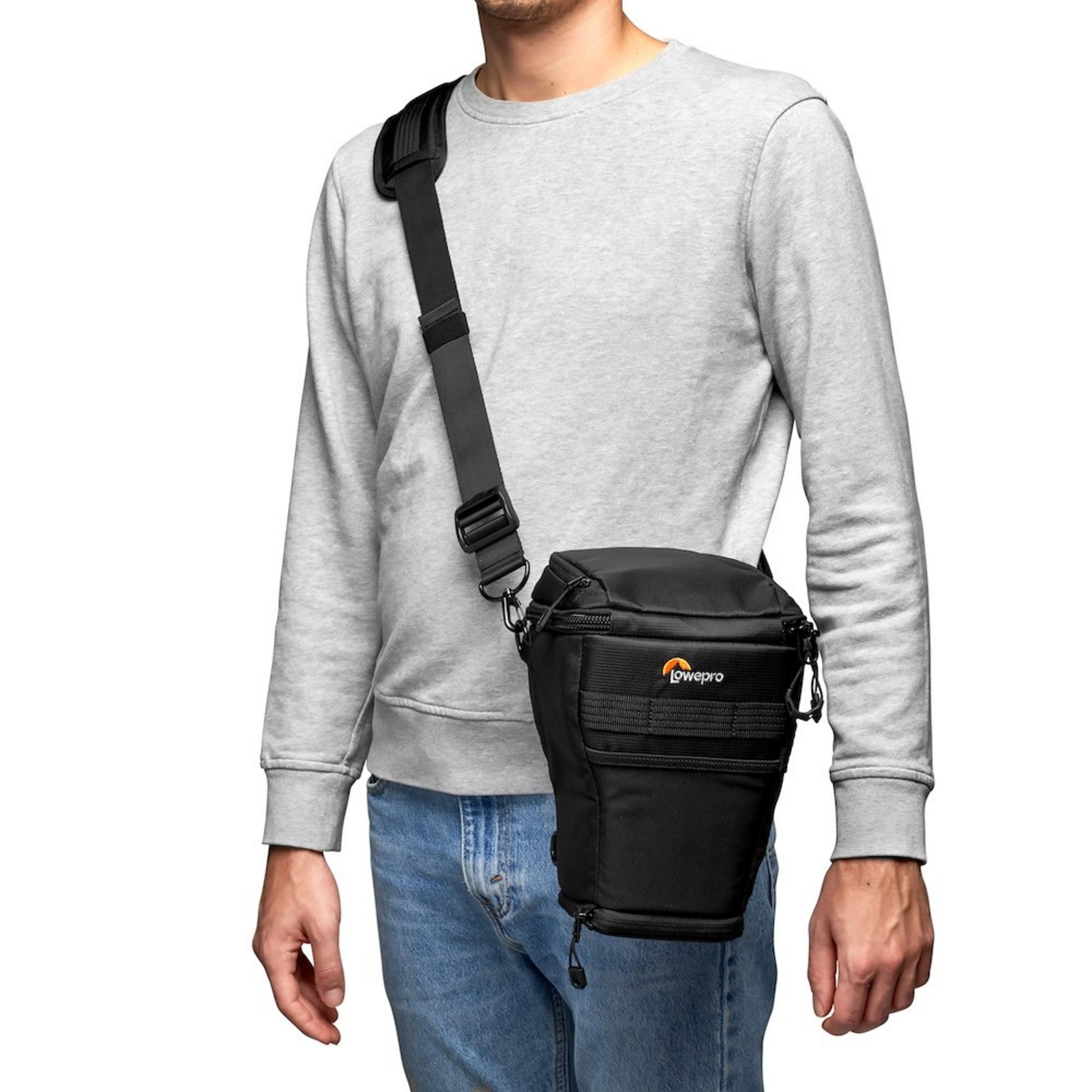 Eine Person trägt eine schwarze Kameratasche über der Schulter. Die Tasche hat einen orangefarbenen Logo-Aufnäher.