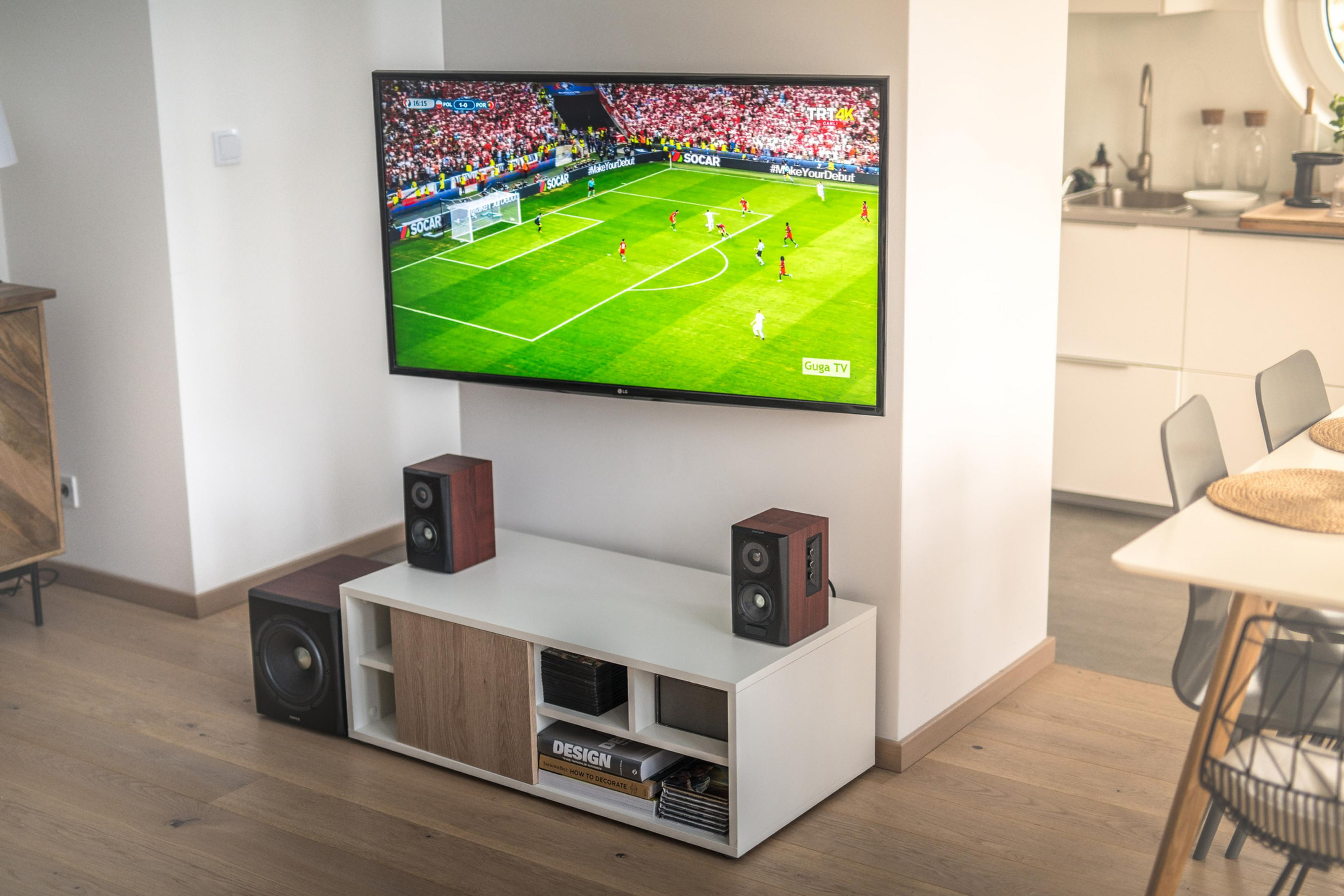Ein Fernseher zeigt ein Fußballspiel. Ein weißes Sideboard mit Lautsprechern steht darunter.