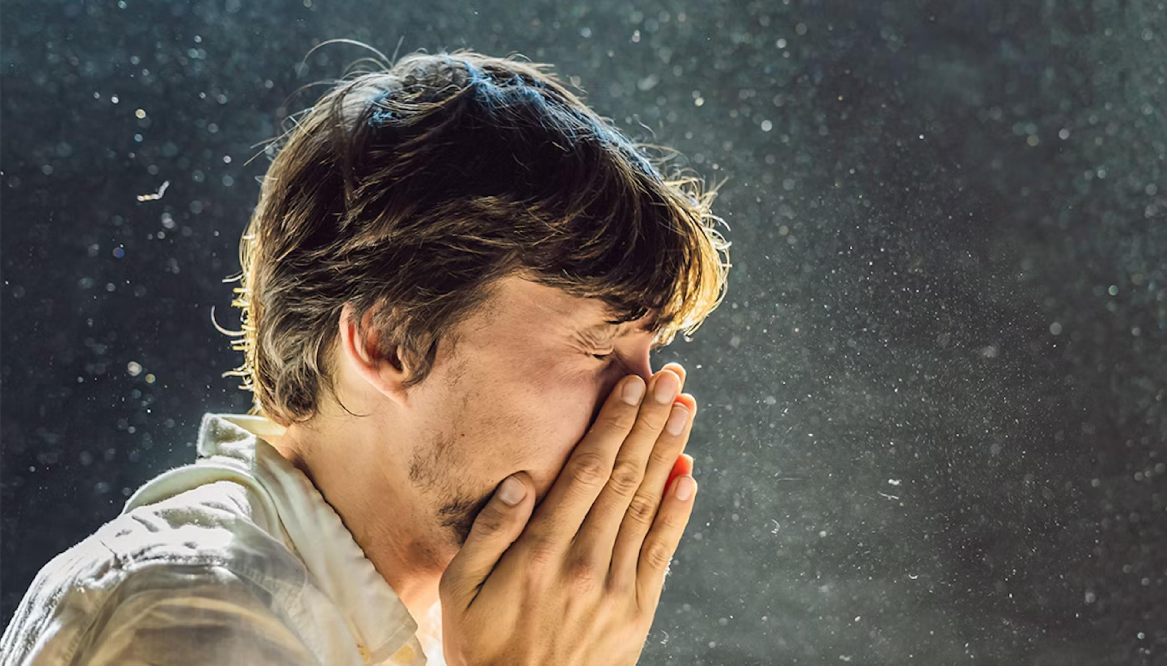 Man sneezes into his hands, surrounded by airborne dust particles.
