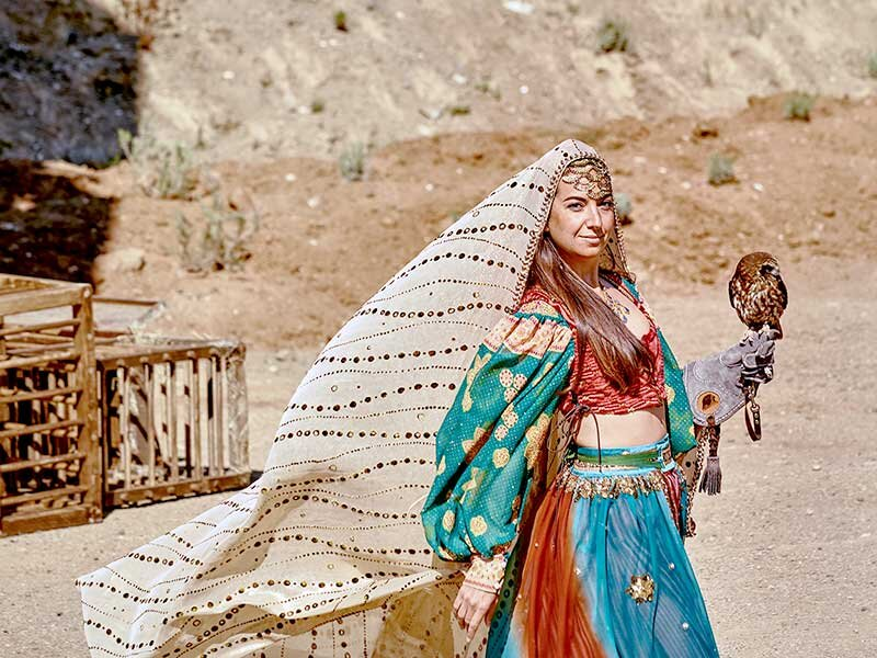 Una mujer con atuendo tradicional sostiene un halcón en la mano. Paisaje desértico al fondo.