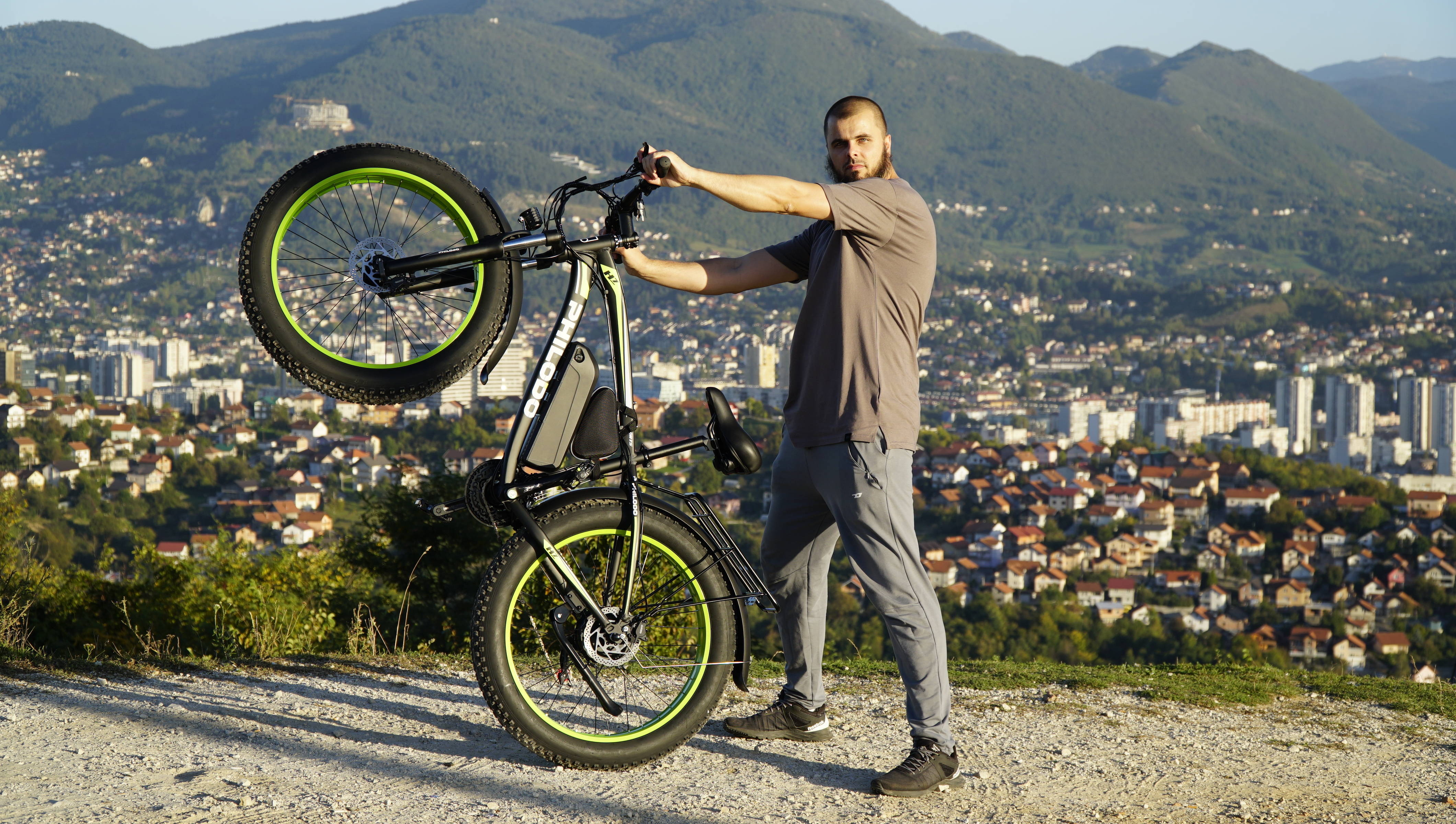 Mann hält ein E-Bike mit grünen Felgen, Blick auf Stadt und Berge.