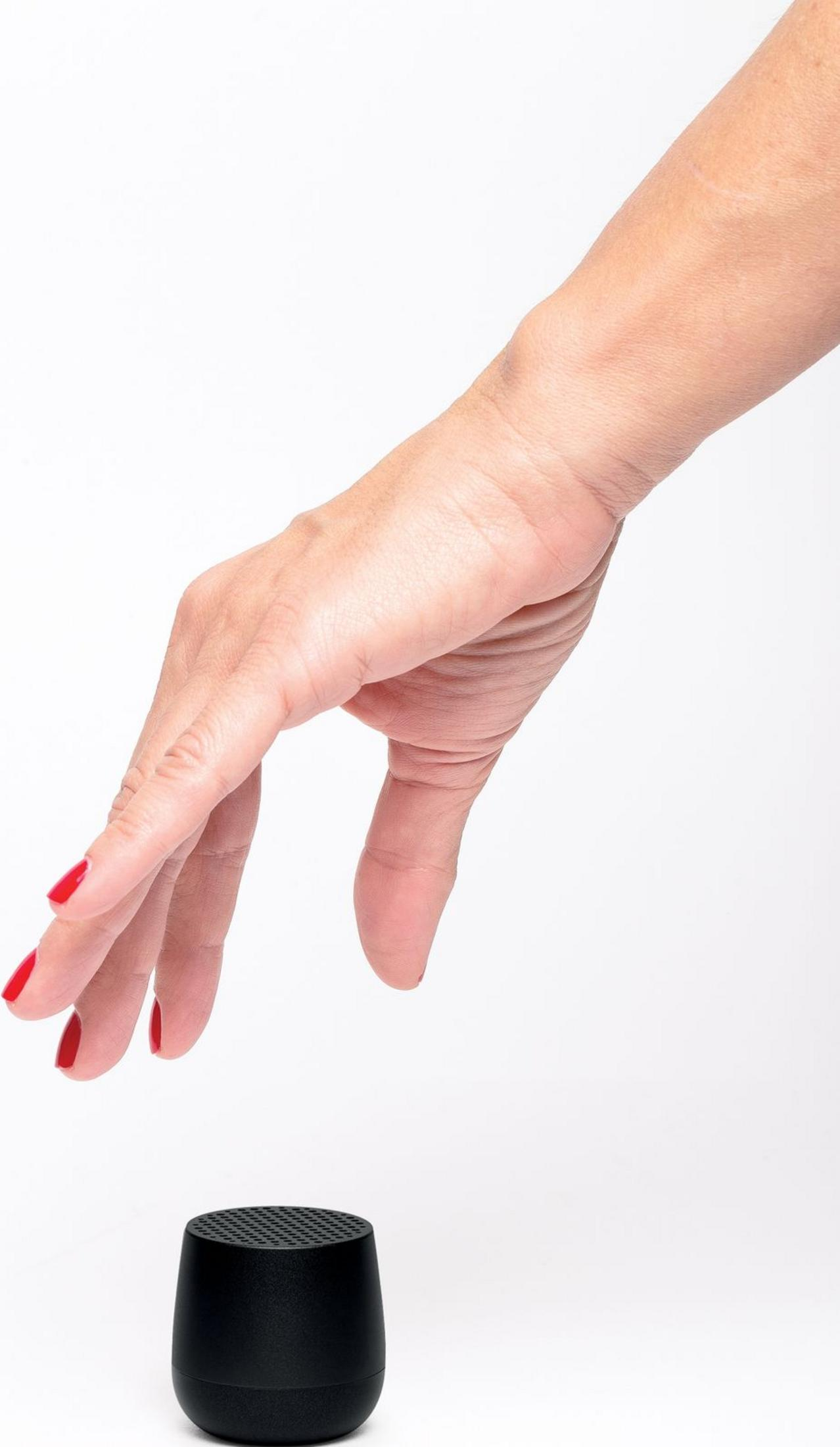 Una mano con unghie rosse si protende verso un piccolo altoparlante nero su sfondo bianco.