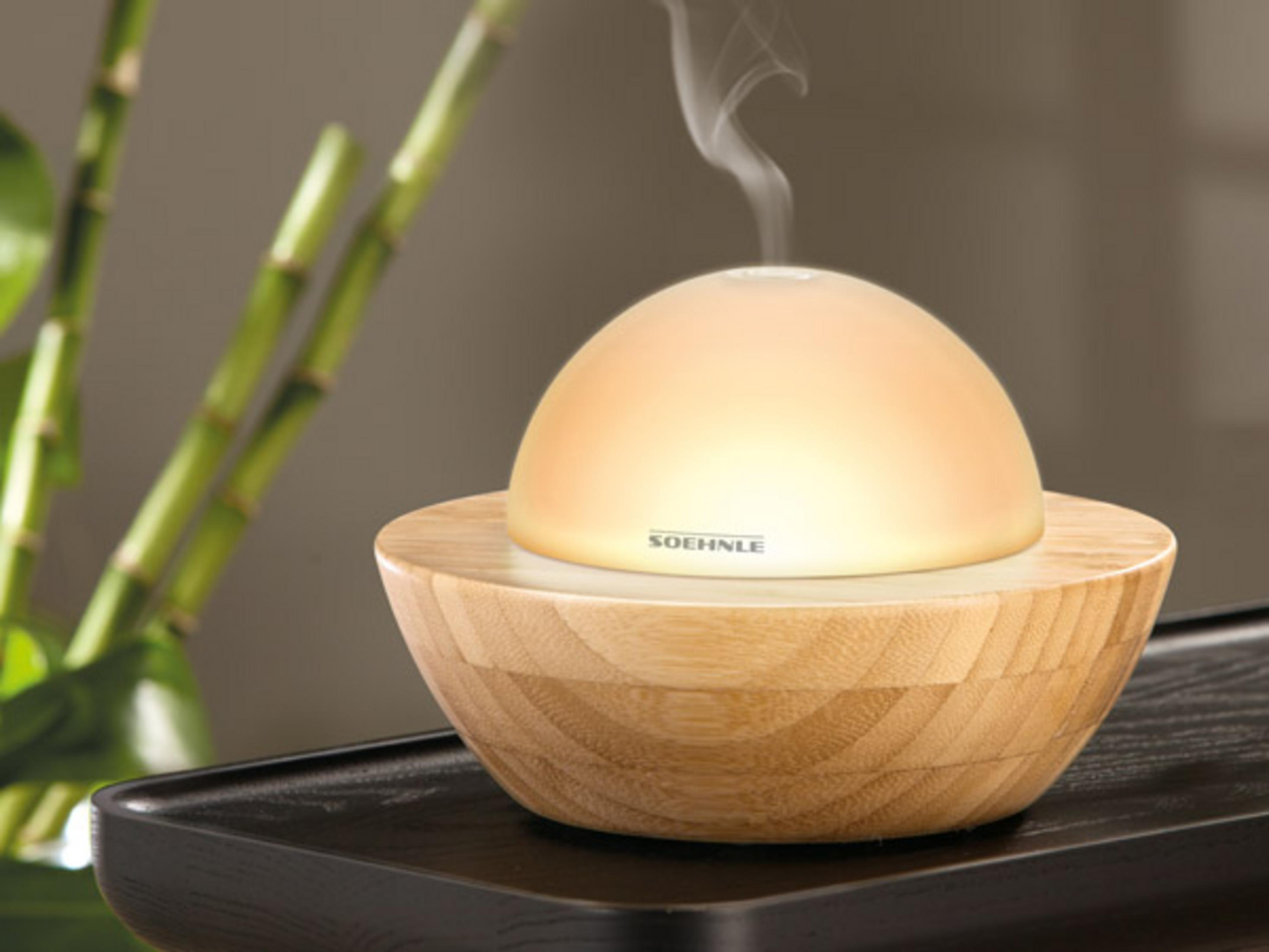 Un diffusore con base in legno e cupola luminosa emette vapore. Bambù sullo sfondo.