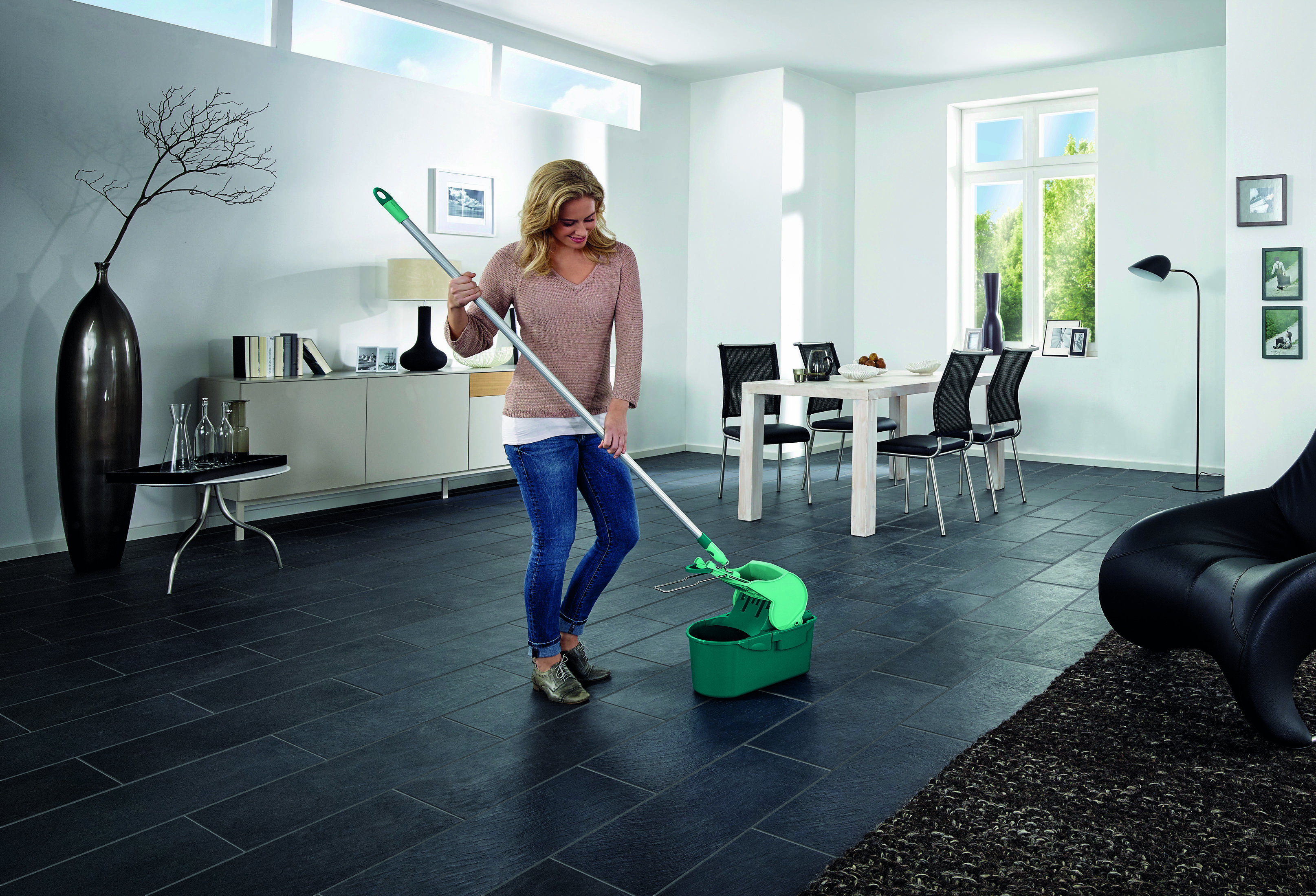 Kobieta w jeansach i swetrze pcha zielony mop z wiadrem.