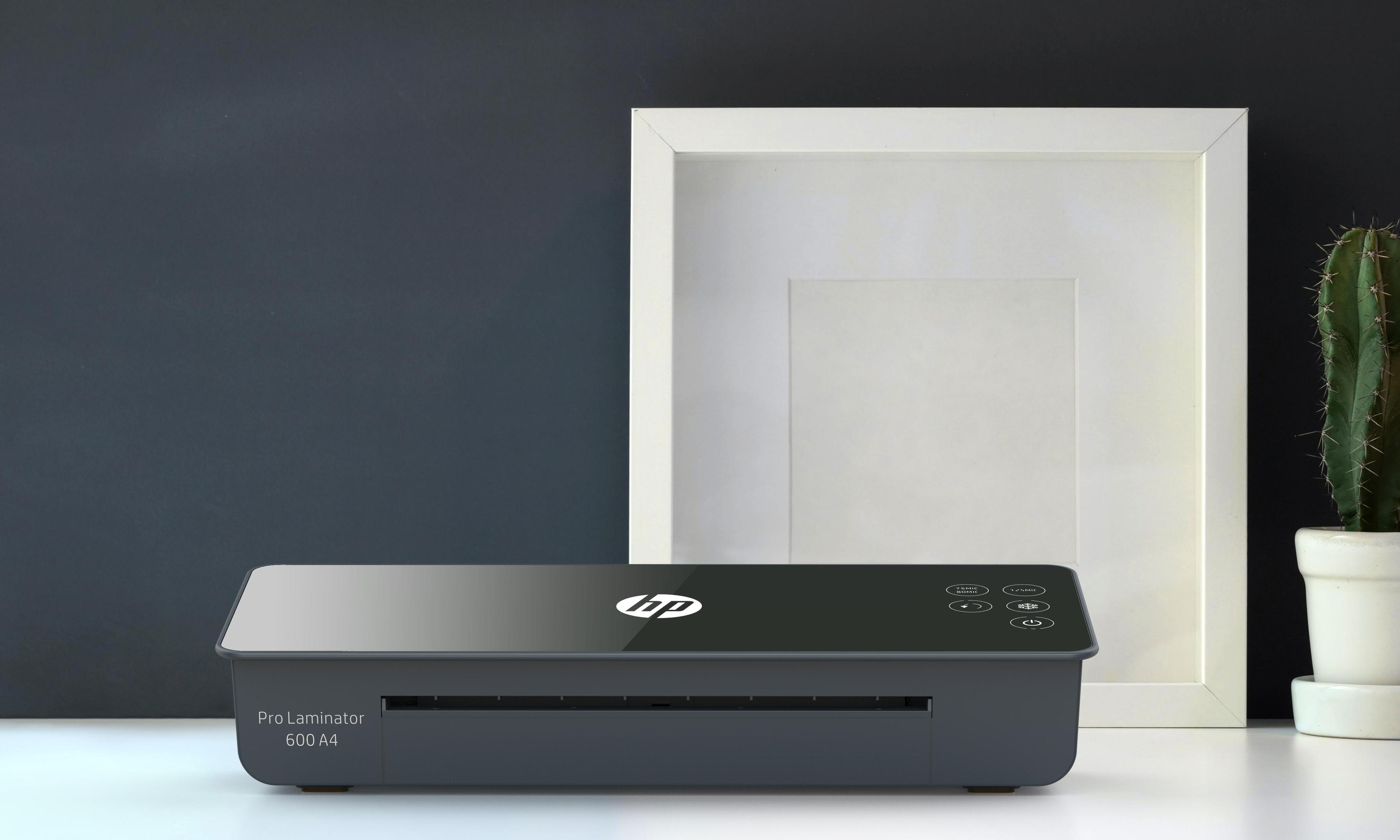Ein dunkler Laminator mit HP-Logo steht auf einem weißen Tisch neben einem weißen Bilderrahmen und einer Topfpflanze.