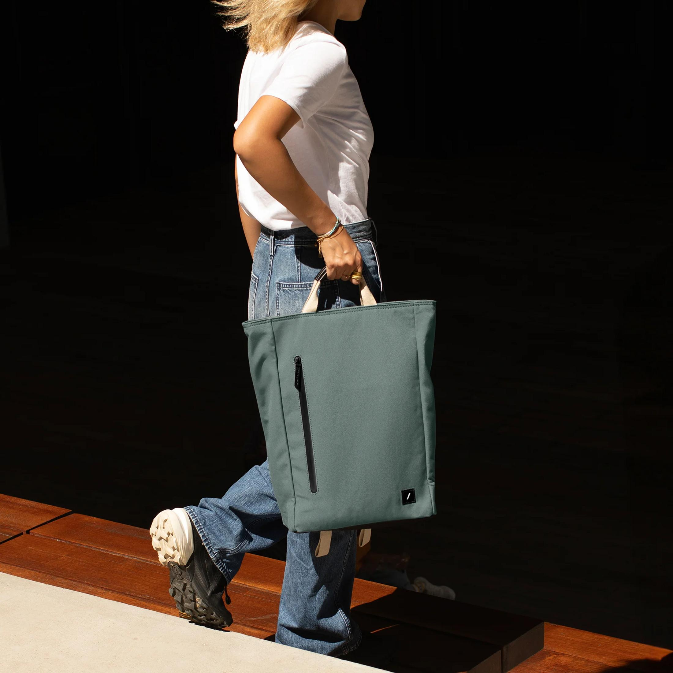 Eine Frau in Jeans und weißem T-Shirt trägt eine tealfarbene Tragetasche mit Reißverschluss.