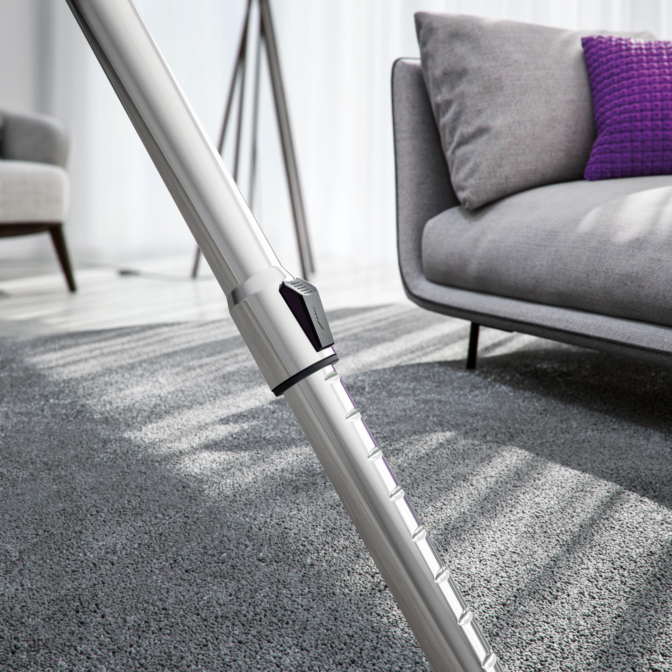 Tubo plateado de aspiradora sobre alfombra gris delante de sofá y lámpara.