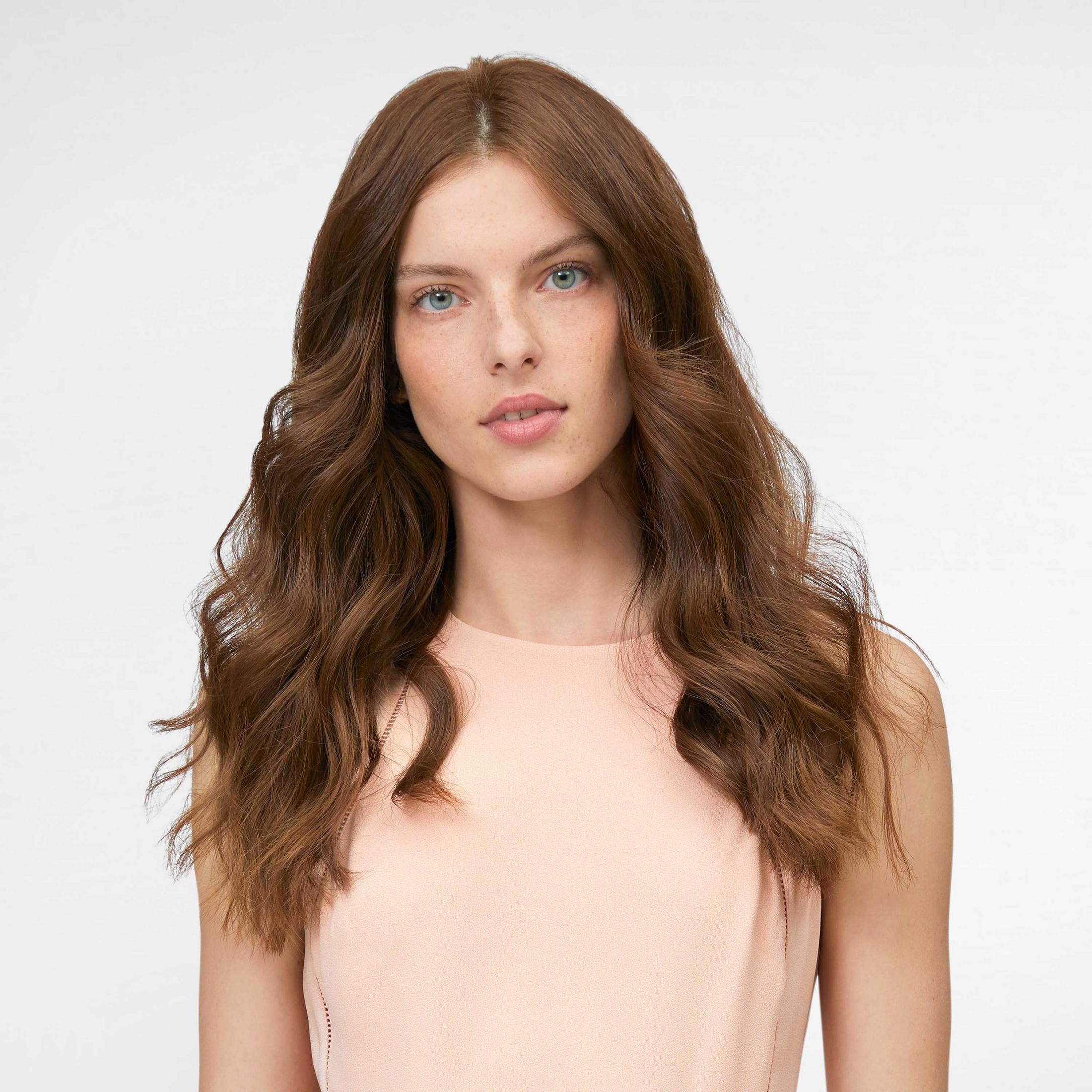 Una donna con lunghi capelli castani e occhi azzurri indossa un abito rosa chiaro.