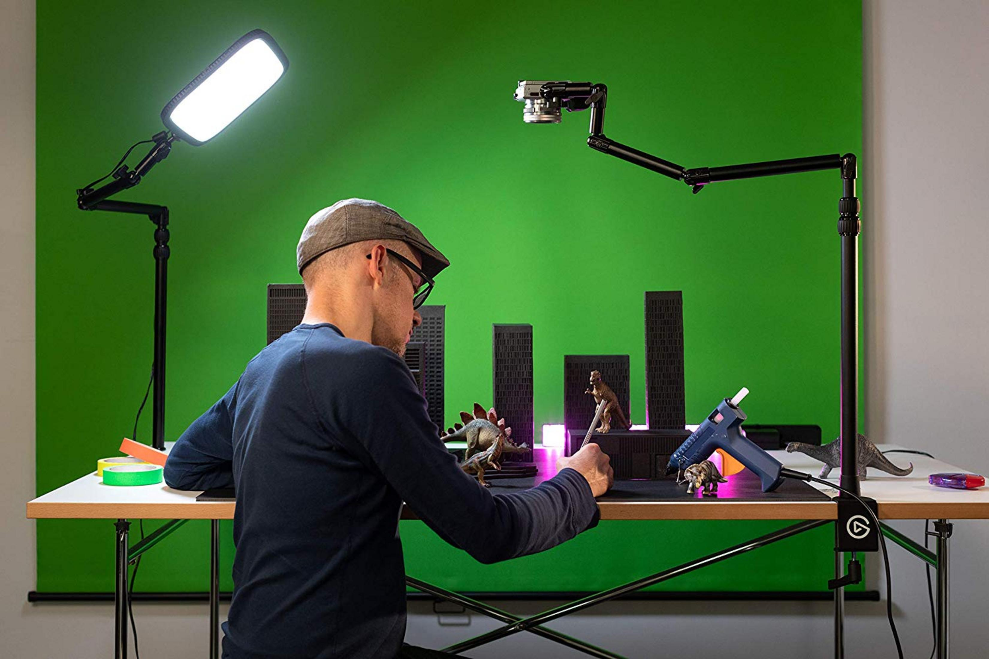 Ein Mann mit Kappe arbeitet an einem Tisch mit Dinosaurierspielzeug, Stadtmodellen und einer Heißklebepistole.