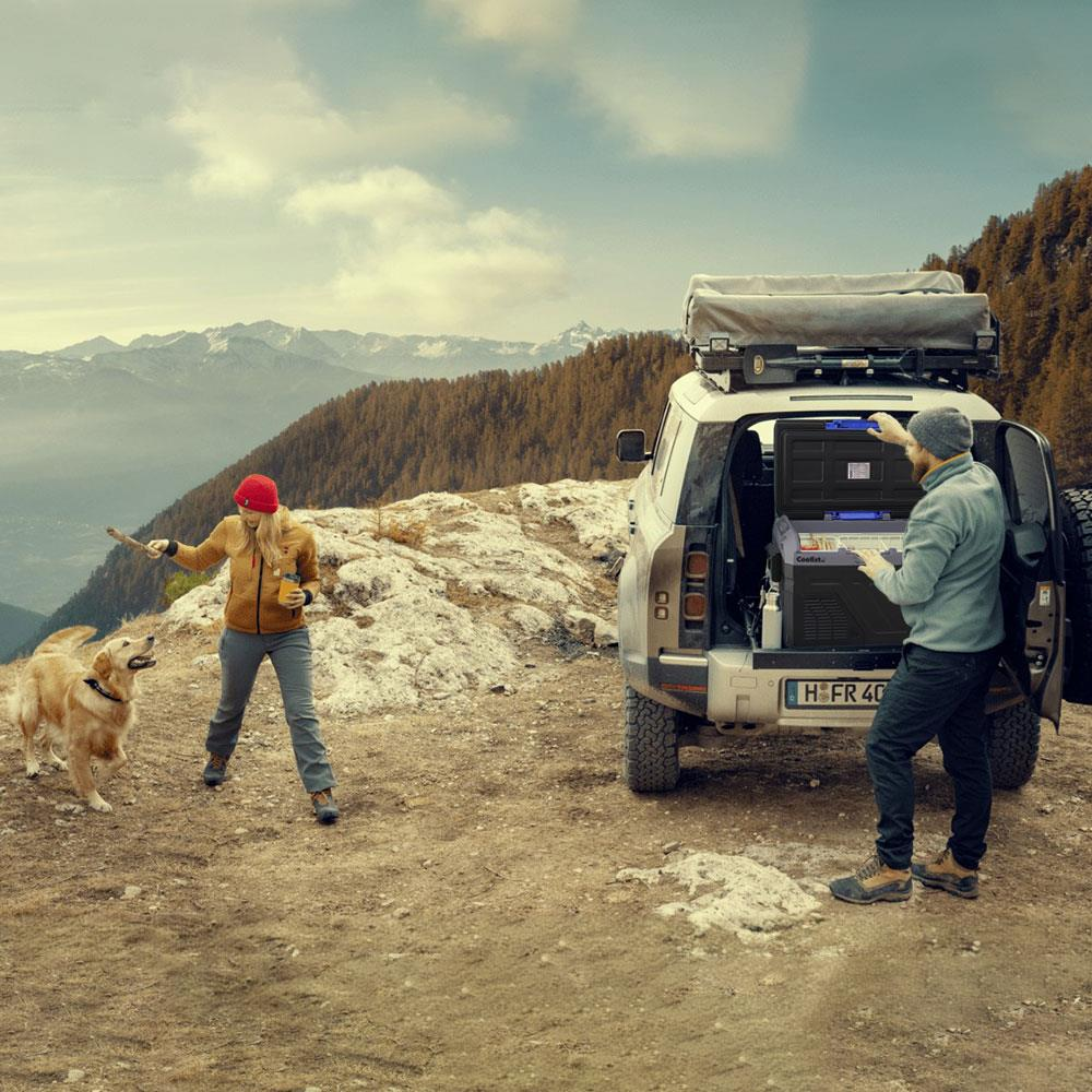 Adam SUV'a soğutucu yüklüyor, kadın dağ manzarasında köpekle oynuyor.