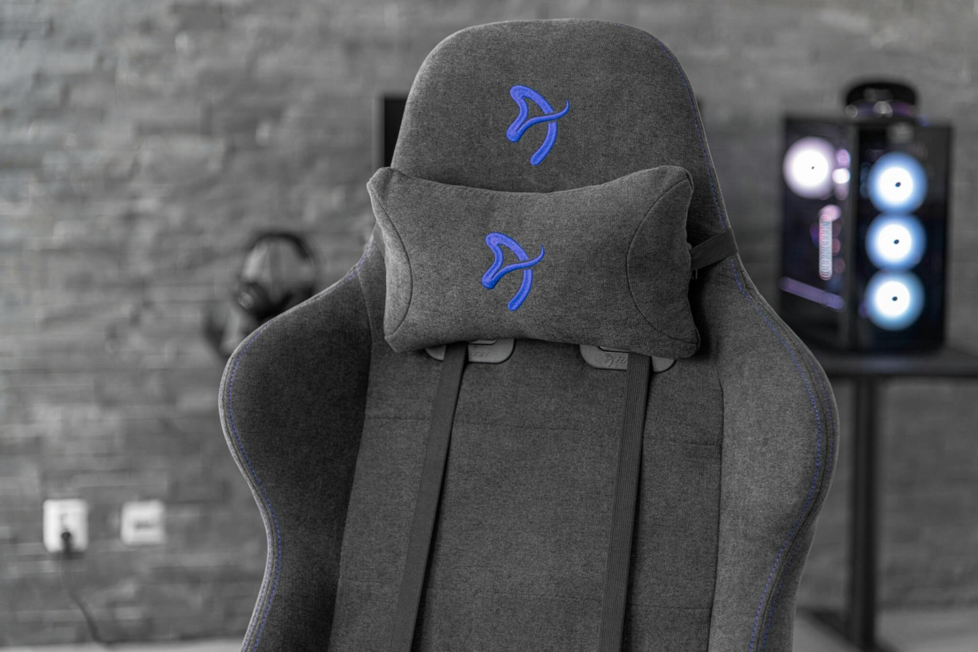 Silla gamer gris con costuras y logo azules, con un PC cerca.