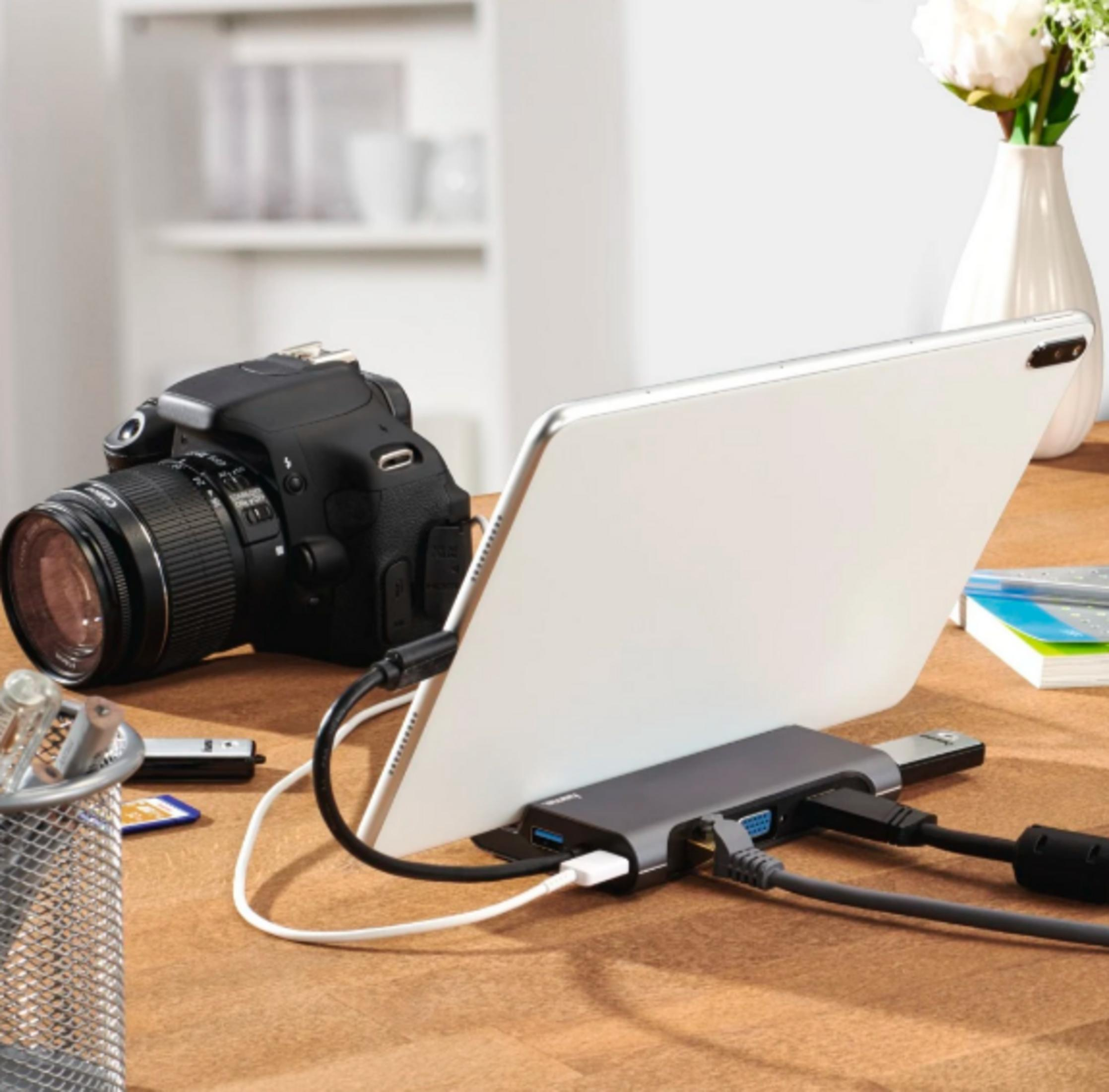 Un laptop, una fotocamera, un hub USB e un portapenne su una scrivania di legno.
