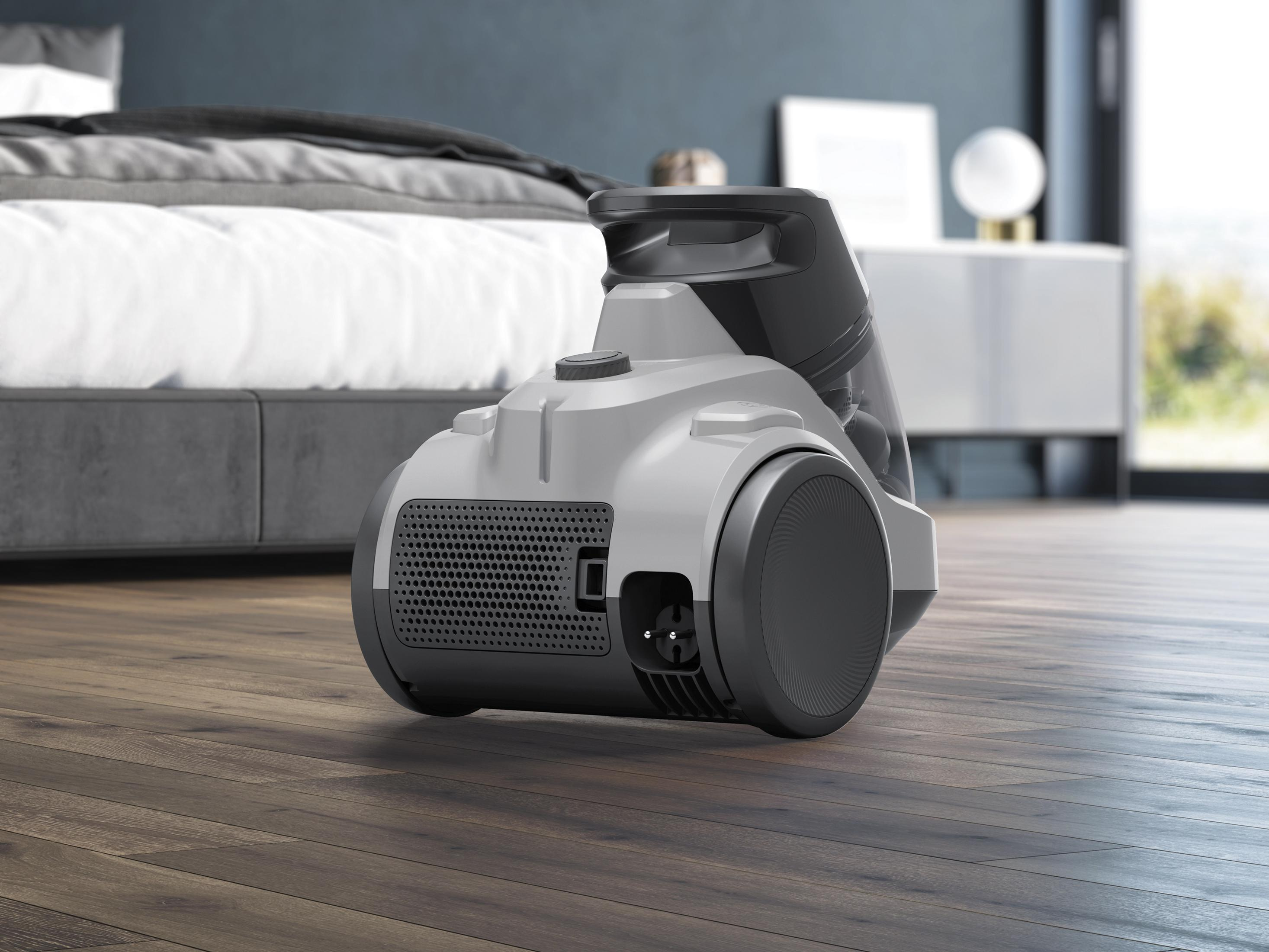 Aspiradora gris sobre suelo de madera junto a una cama.