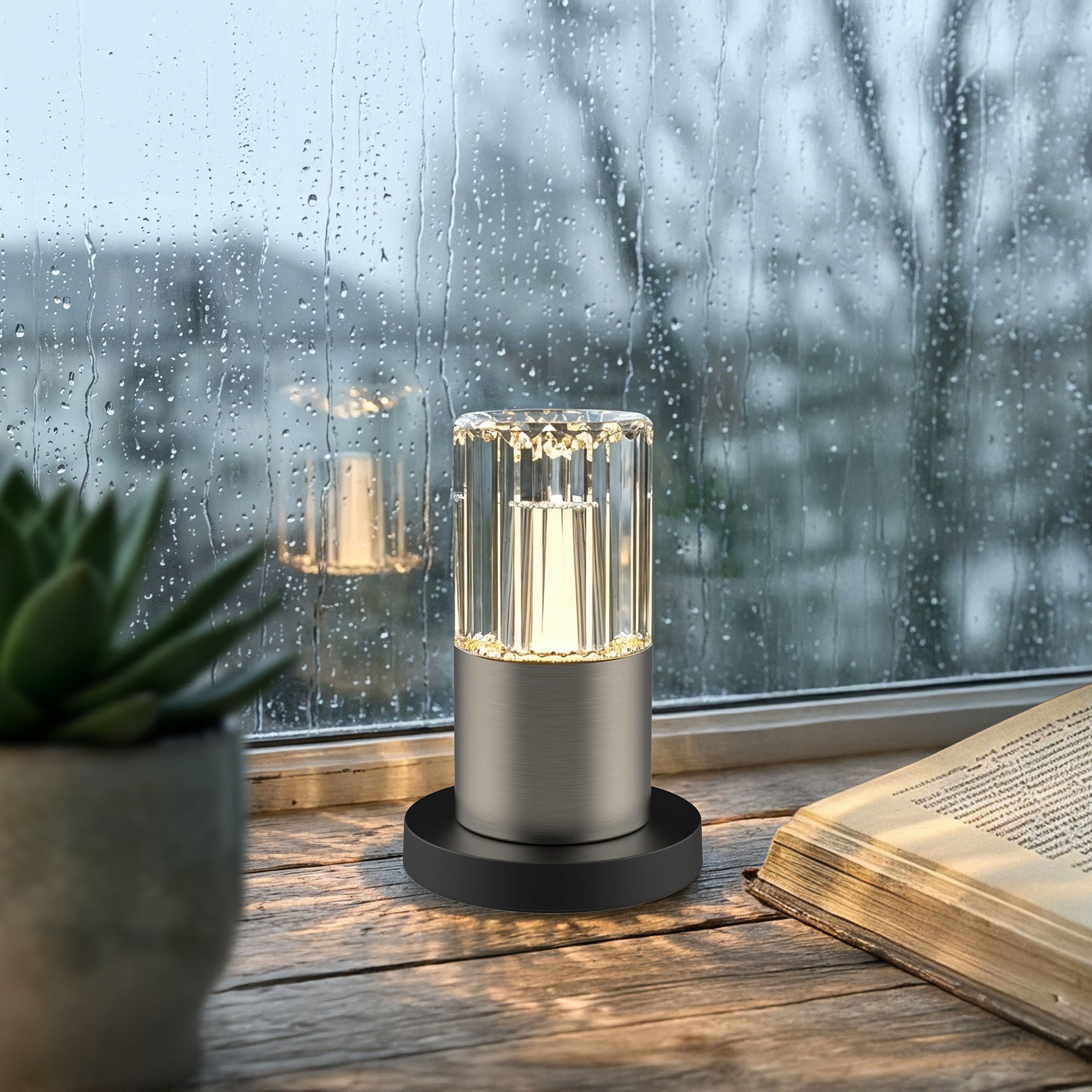 Kristalllampe auf Holztisch vor regennassem Fenster, daneben Buch und Pflanze.