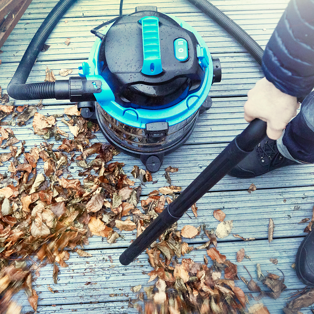 Ein Mann saugt mit einem blauen und schwarzen Nass-/Trockensauger trockene Blätter von einer Holzterrasse.
