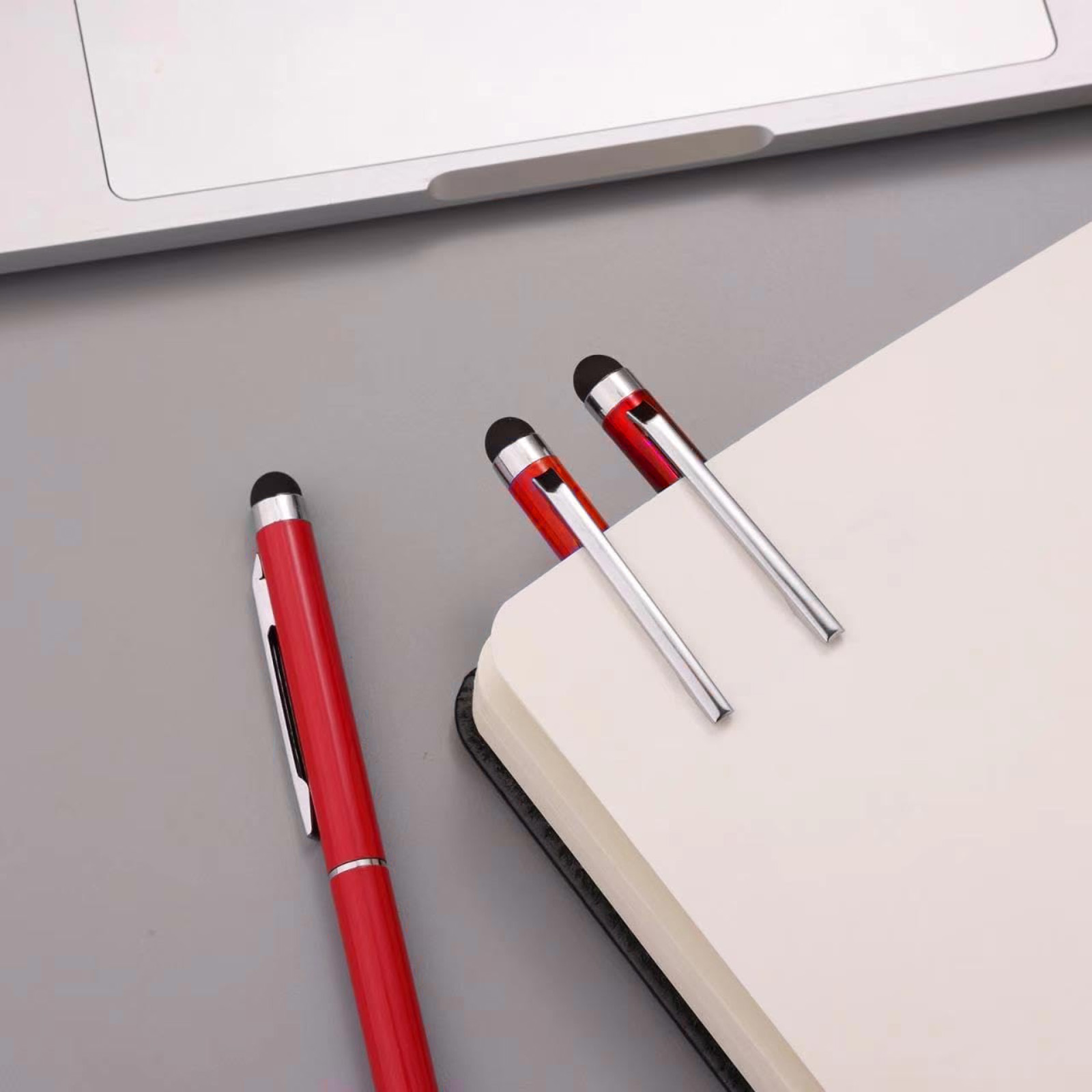 Ein roter Stift liegt links neben einem Notizbuch mit zwei silbernen Stiften.