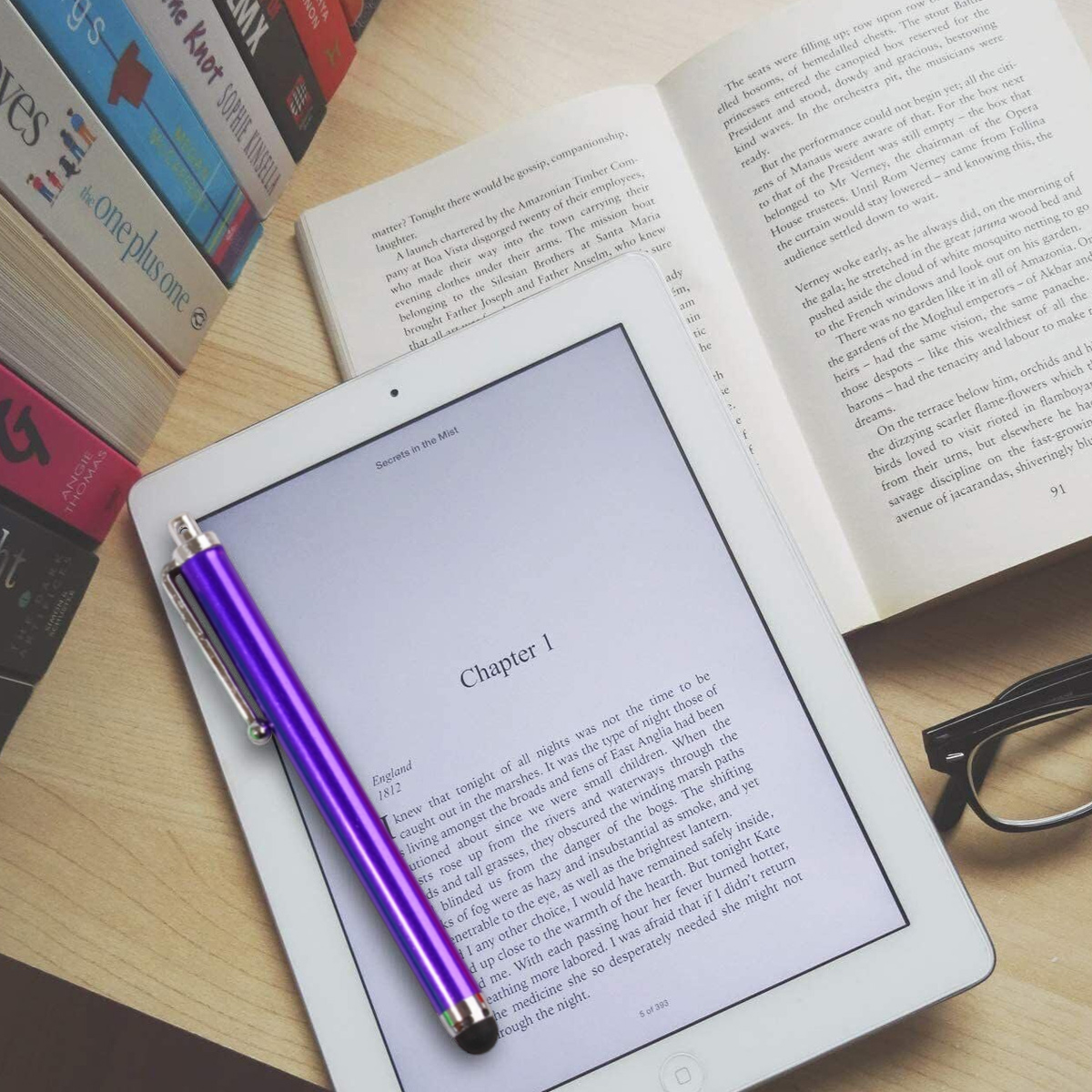 Ein offenes Buch und ein Tablet liegen auf einem Holztisch, daneben Stifte und Brille.