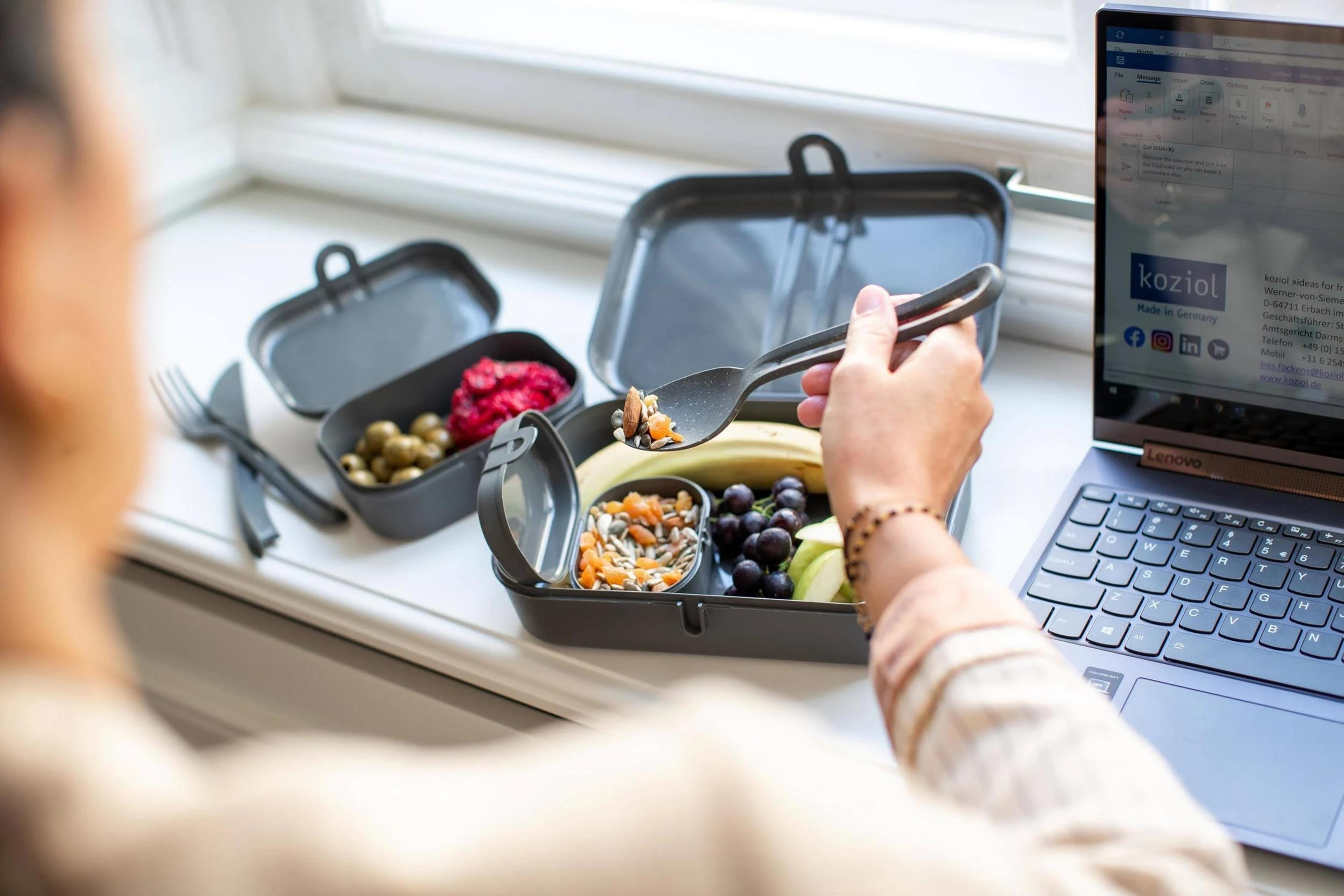 Persona mangia da una scatola per il pranzo con frutta, noci e semi accanto a un laptop.
