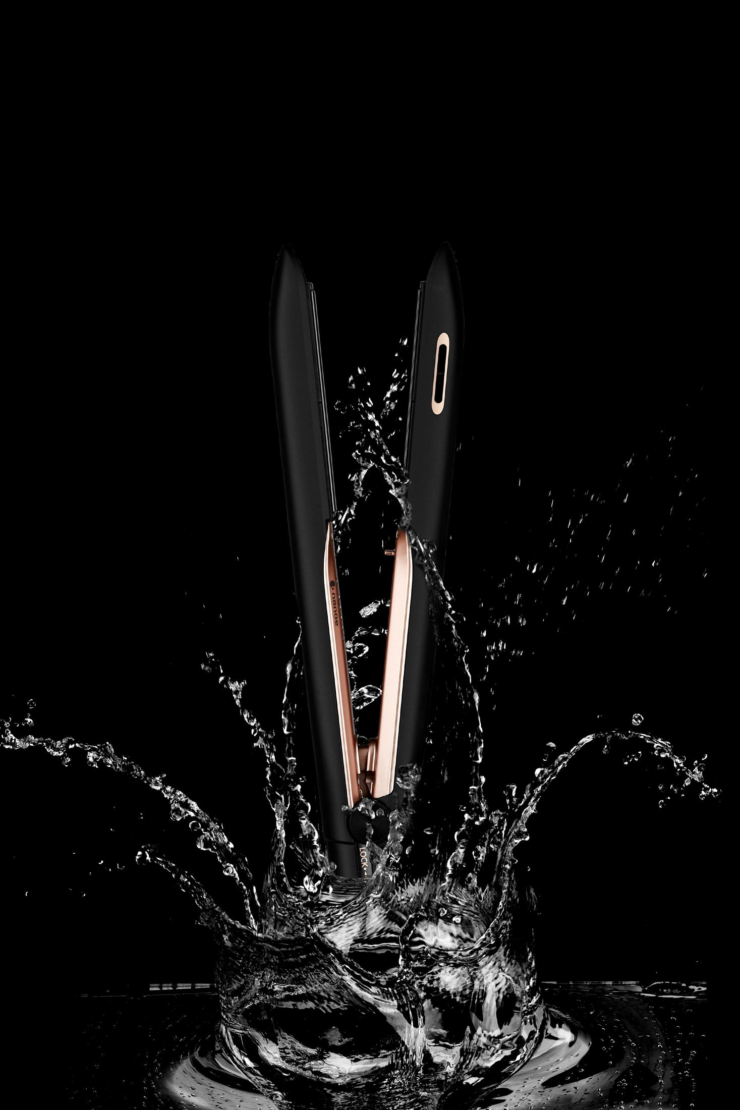 A black and rose gold hair straightener is partially submerged in water, creating splashes.