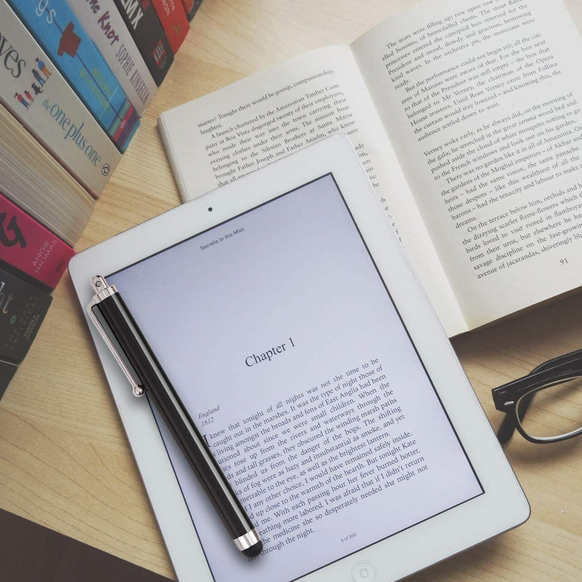 Ein Tablet und ein offenes Buch liegen auf einem Holztisch, daneben ein Stift und Bücher.