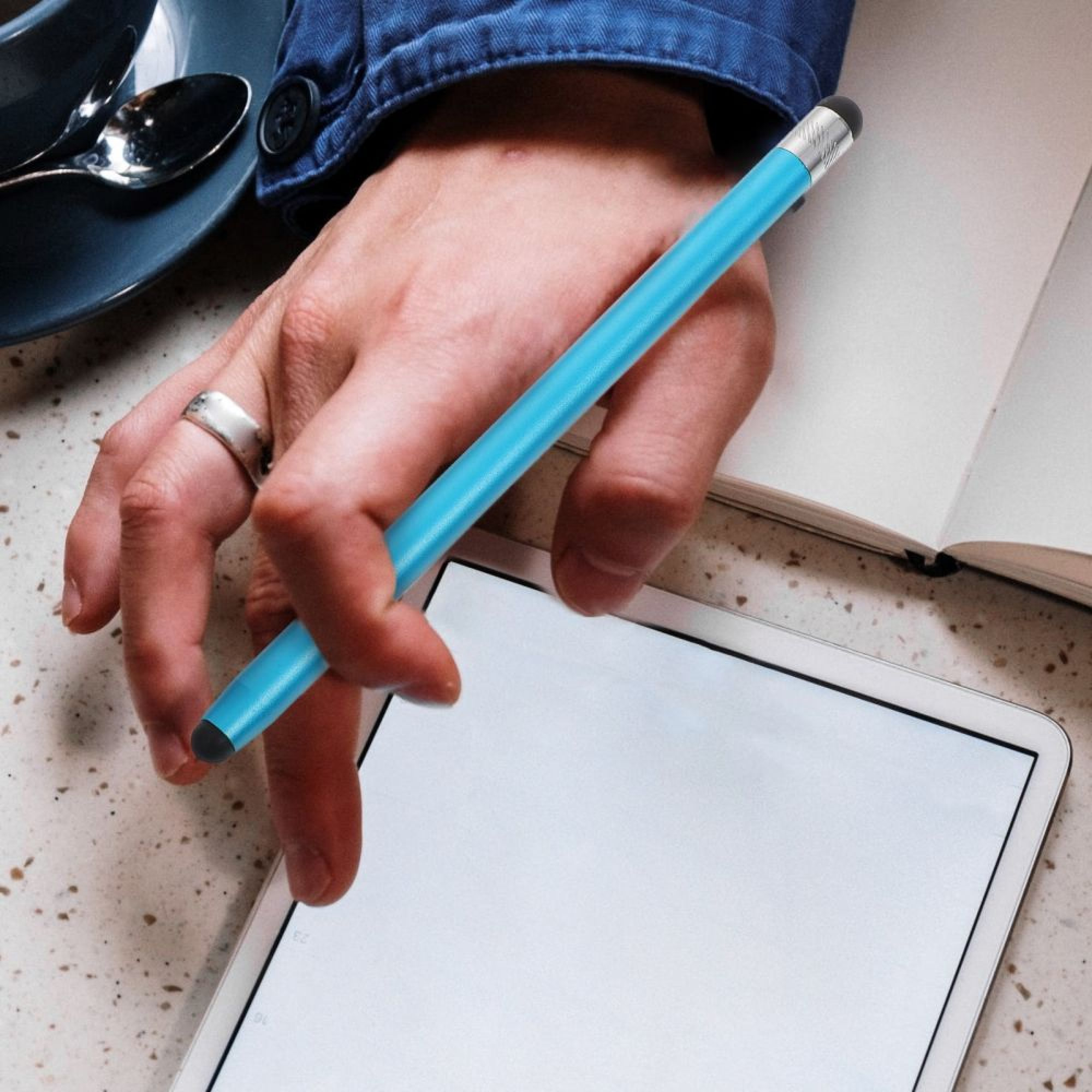Eine Hand hält einen blauen Stift über einem Tablet neben einem Buch.