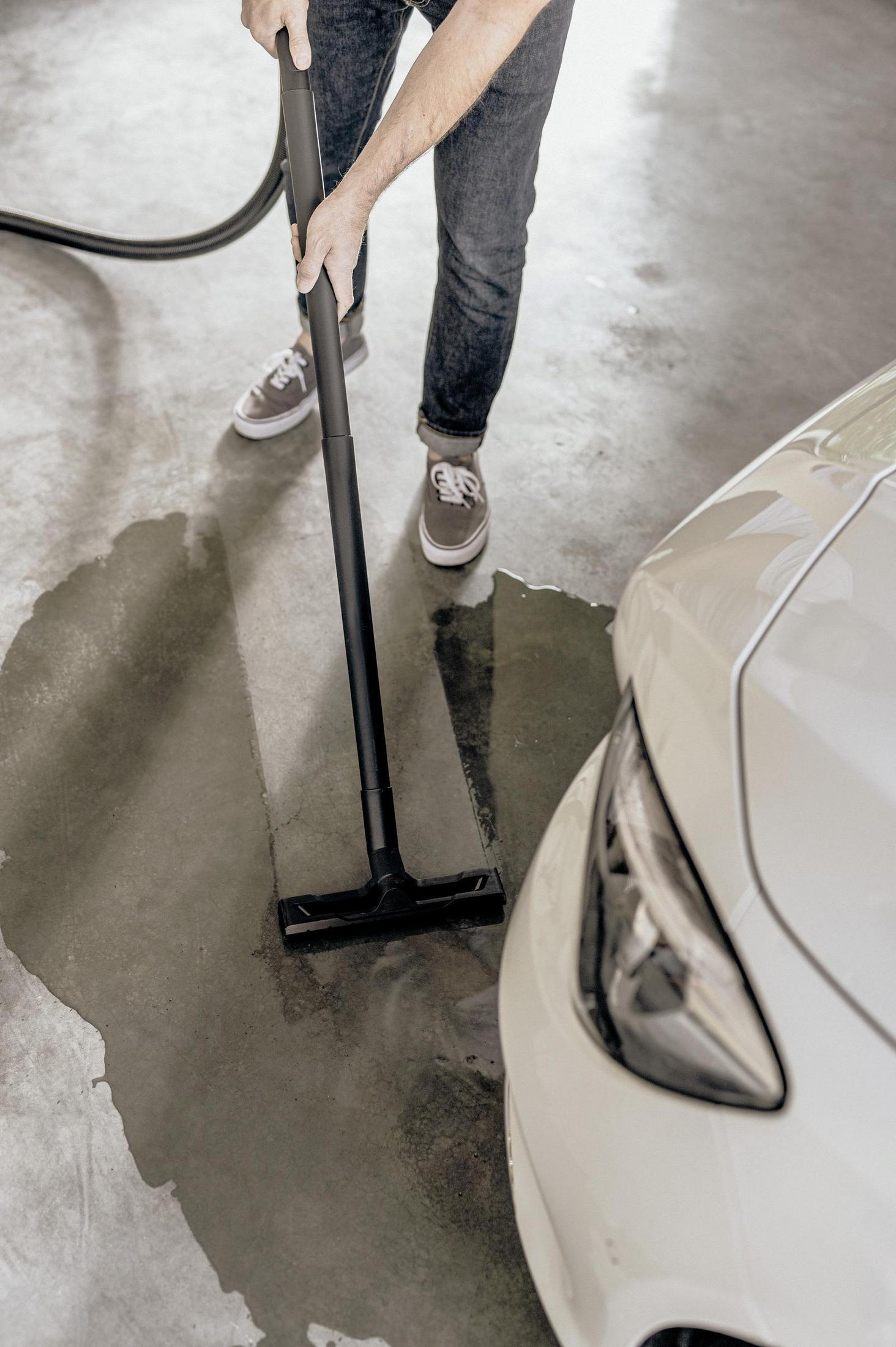 Man vacuums water from concrete floor next to a white car.