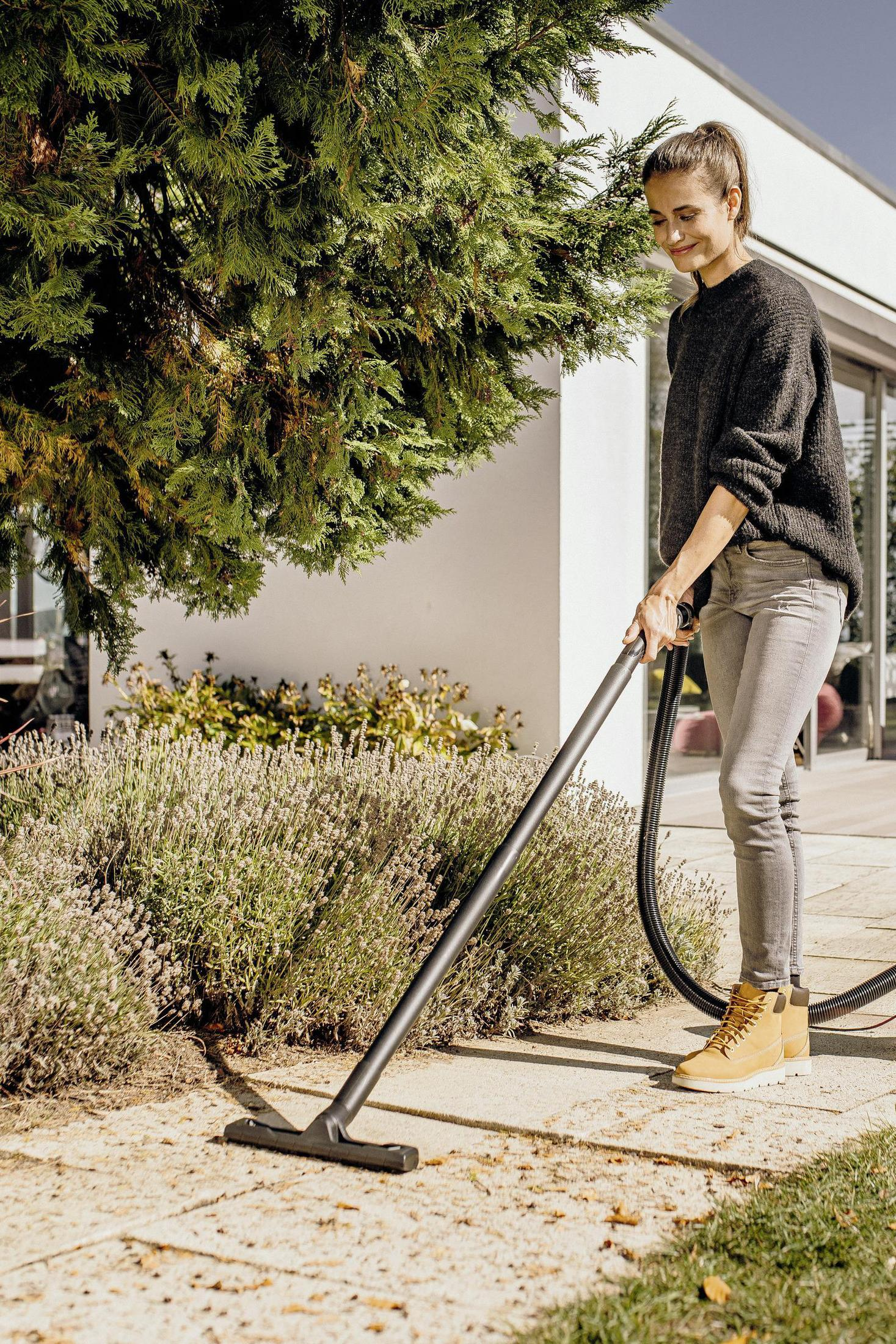 A woman vacuums a patio near lavender bushes and a large evergreen tree.