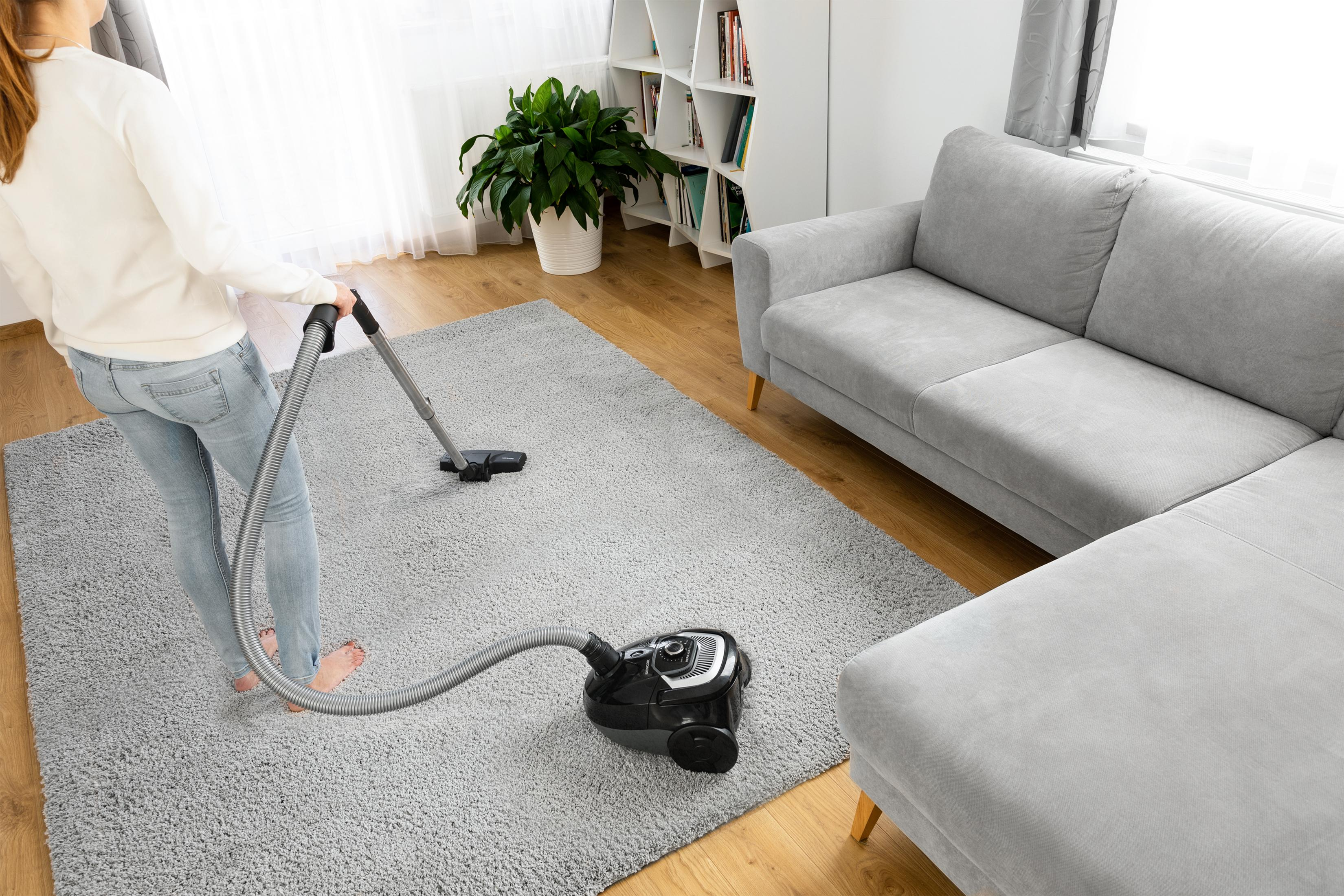 Eine Person saugt einen grauen Teppich in einem Wohnzimmer mit einem Sofa.