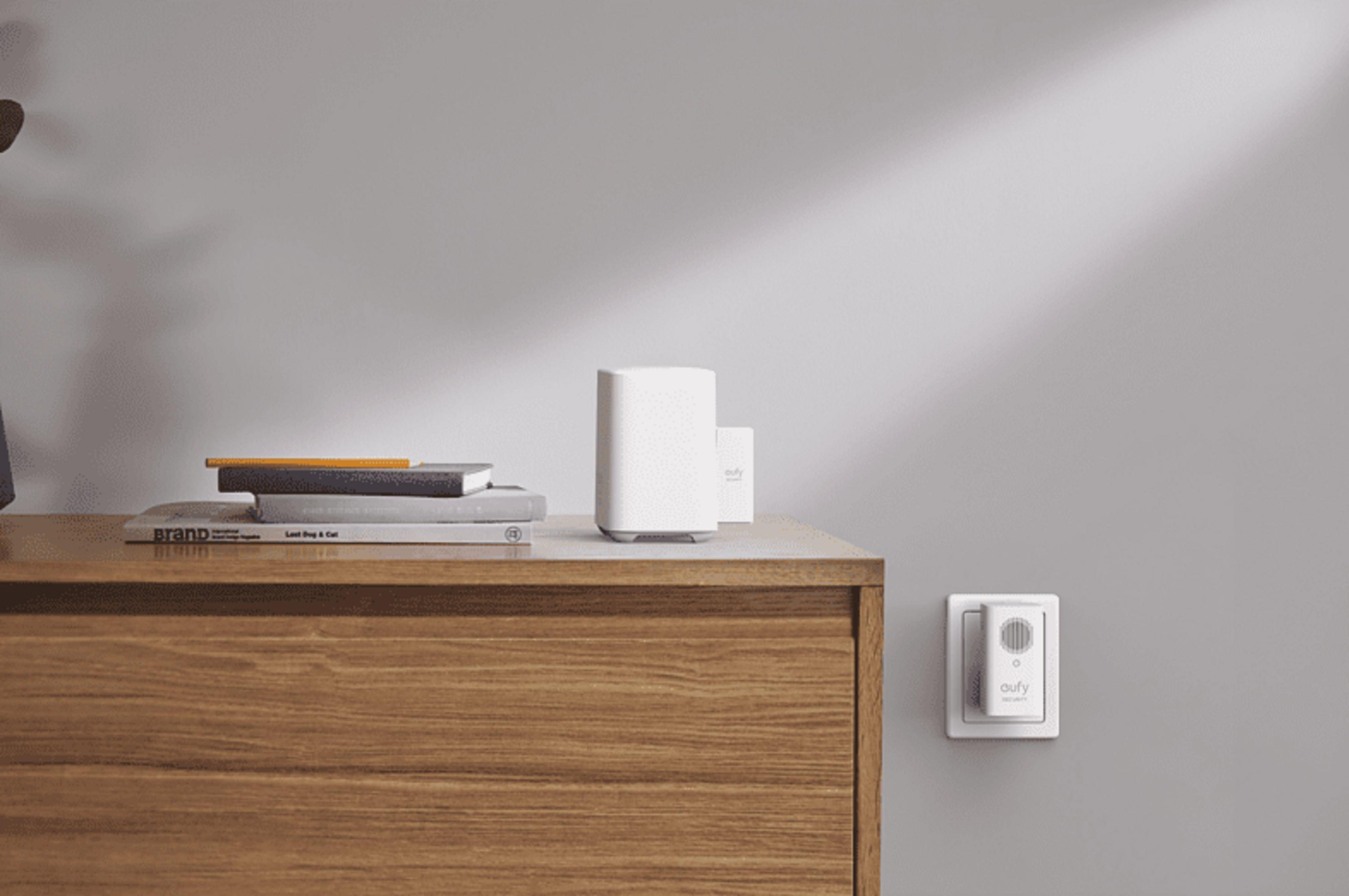 Weißes Eufy-Sicherheitsgerät in Steckdose, neben Büchern und Router auf Holztisch.