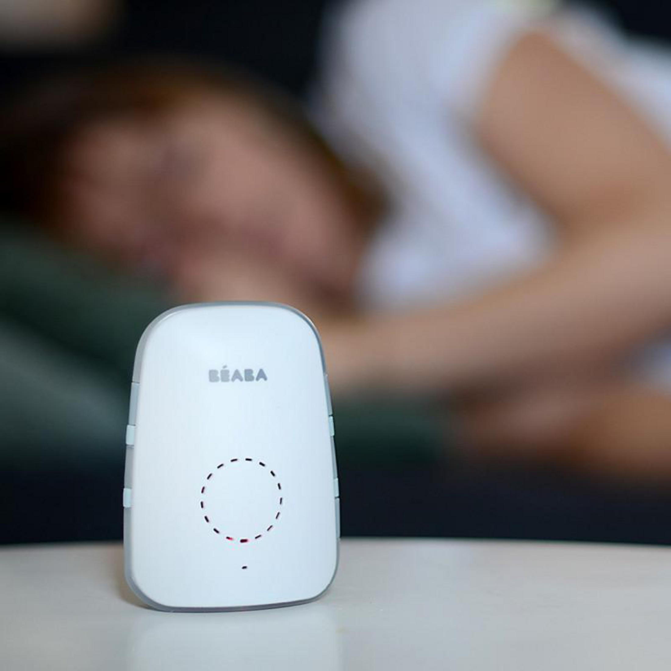 Ein weißer Babyfon-Sender steht auf einem Tisch. Im Hintergrund schläft eine Person.