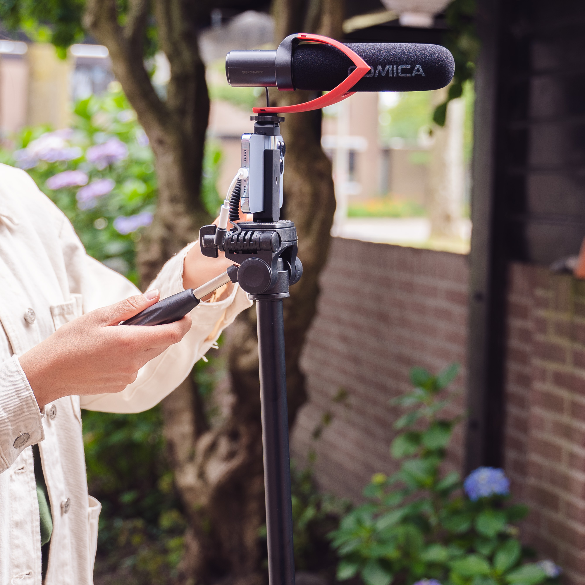 Person holding a tripod with a microphone and camera in a garden.