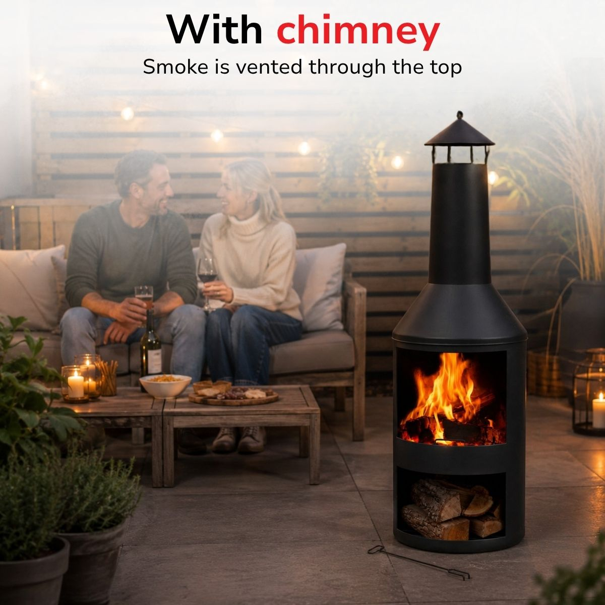 A couple sits on a sofa on a patio, by a lit black chimney with logs stored below.