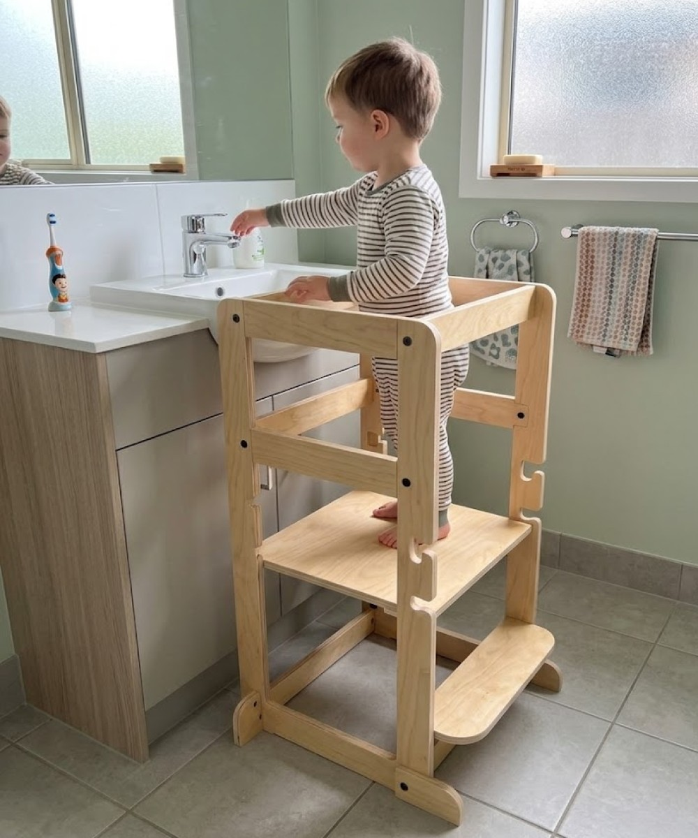 Küçük bir çocuk lavaboda ahşap tabure üzerinde duruyor ve musluğu açıyor.