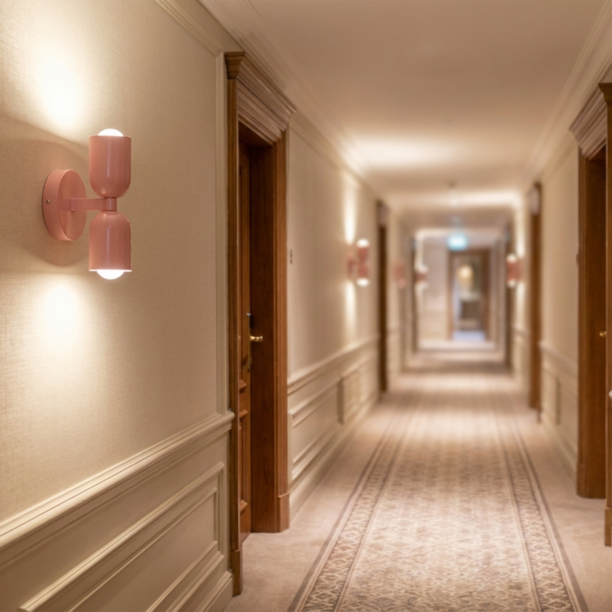 Una lámpara de pared rosa ilumina un largo pasillo de hotel con alfombra y puertas de madera.