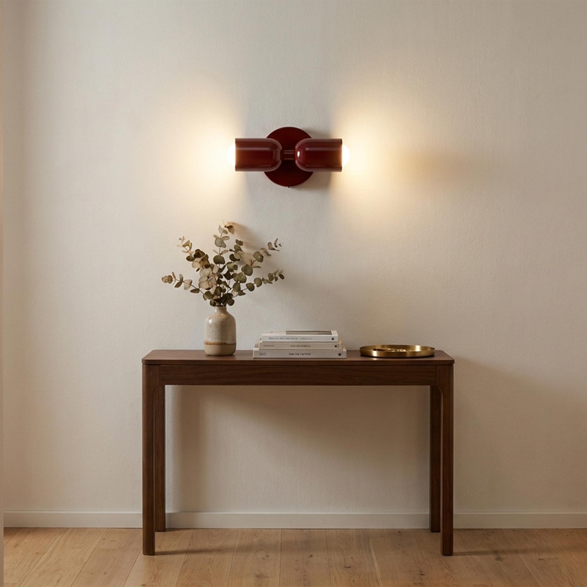 Un aplique de pared rojo ilumina una mesa de madera con jarrón, libros y bandeja.