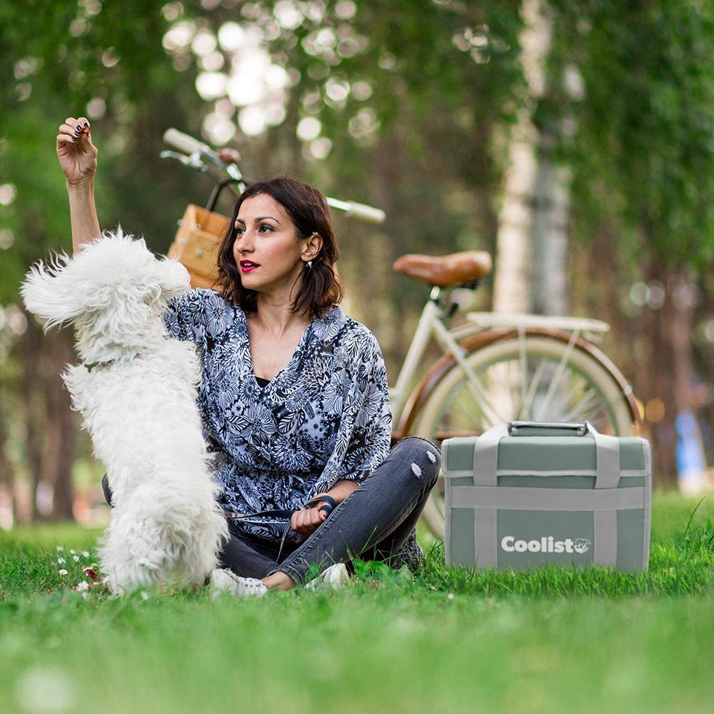 Kadın parkta beyaz köpekle, arkada bisiklet ve soğutucu çanta.