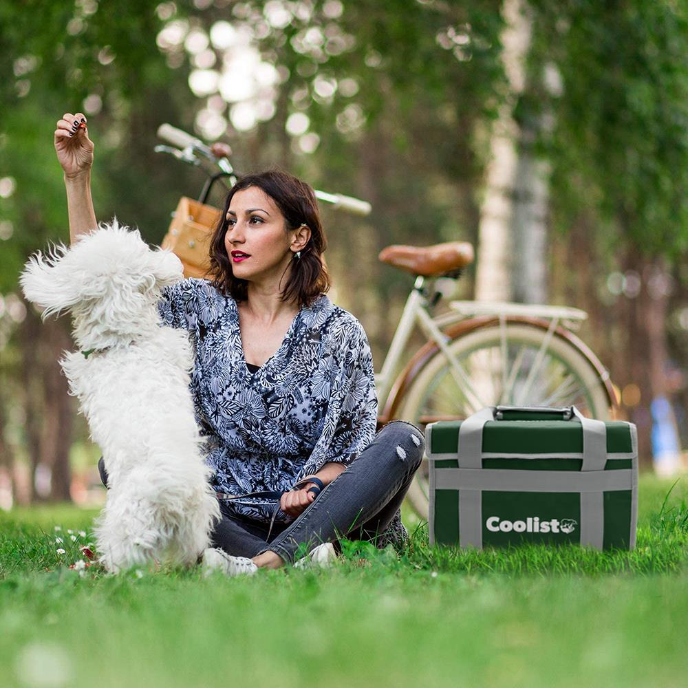 Bir kadın bisiklet ve soğutucu çantasının yanında çimlerde beyaz bir köpekle oynuyor.