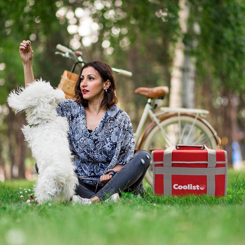 Kadın, bisiklet ve soğutucu çantasının yanında çimlerde beyaz bir köpekle oynuyor.