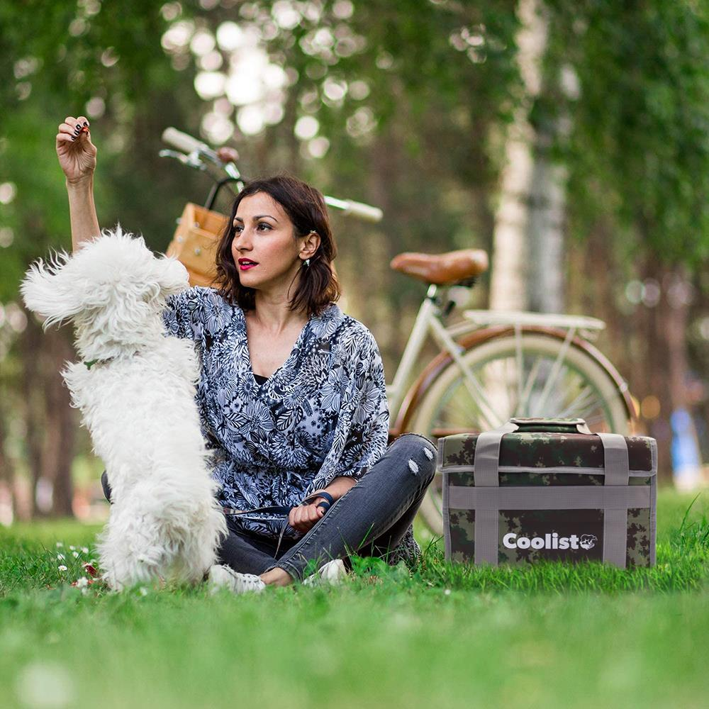Bir kadın çimenlikte, soğutucu çanta ve bisikletin yanında beyaz bir köpekle oynuyor.
